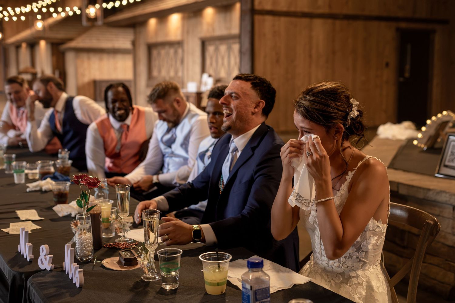 Bride and groom laugh emotionally at sweetheart table during rustic barn wedding reception.