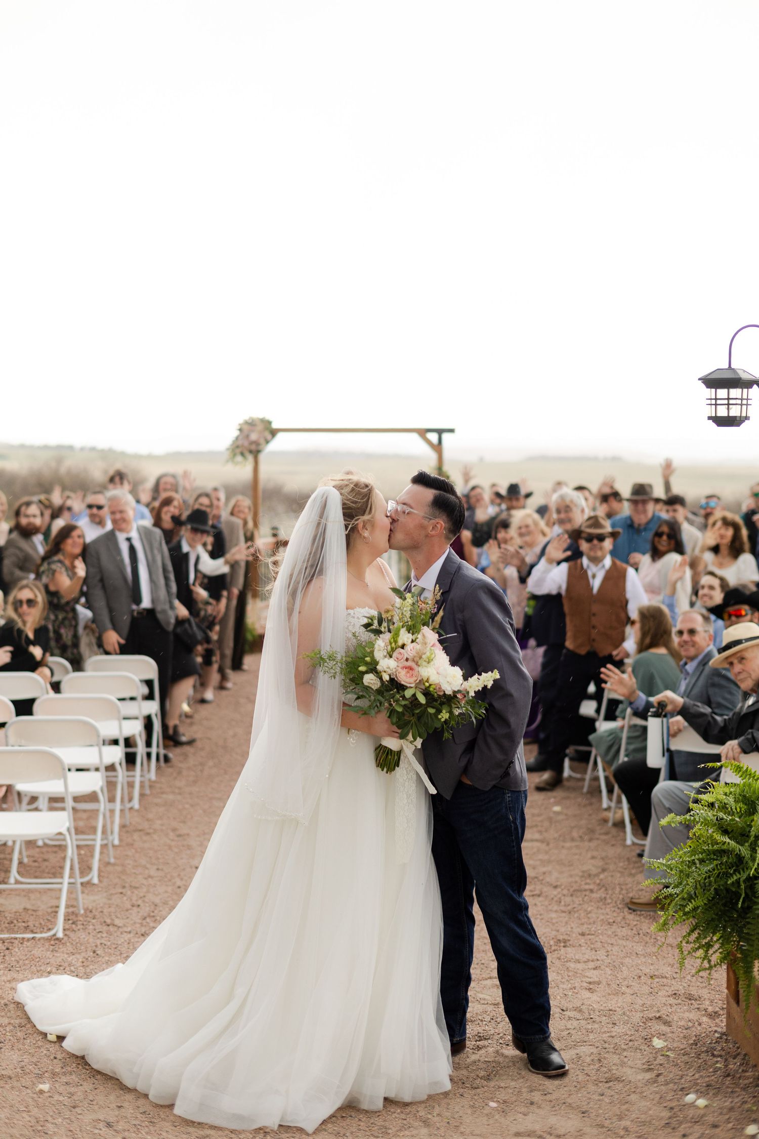 Bride and groom share a first kiss at outdoor desert wedding ceremony with guests cheering along the aisle.