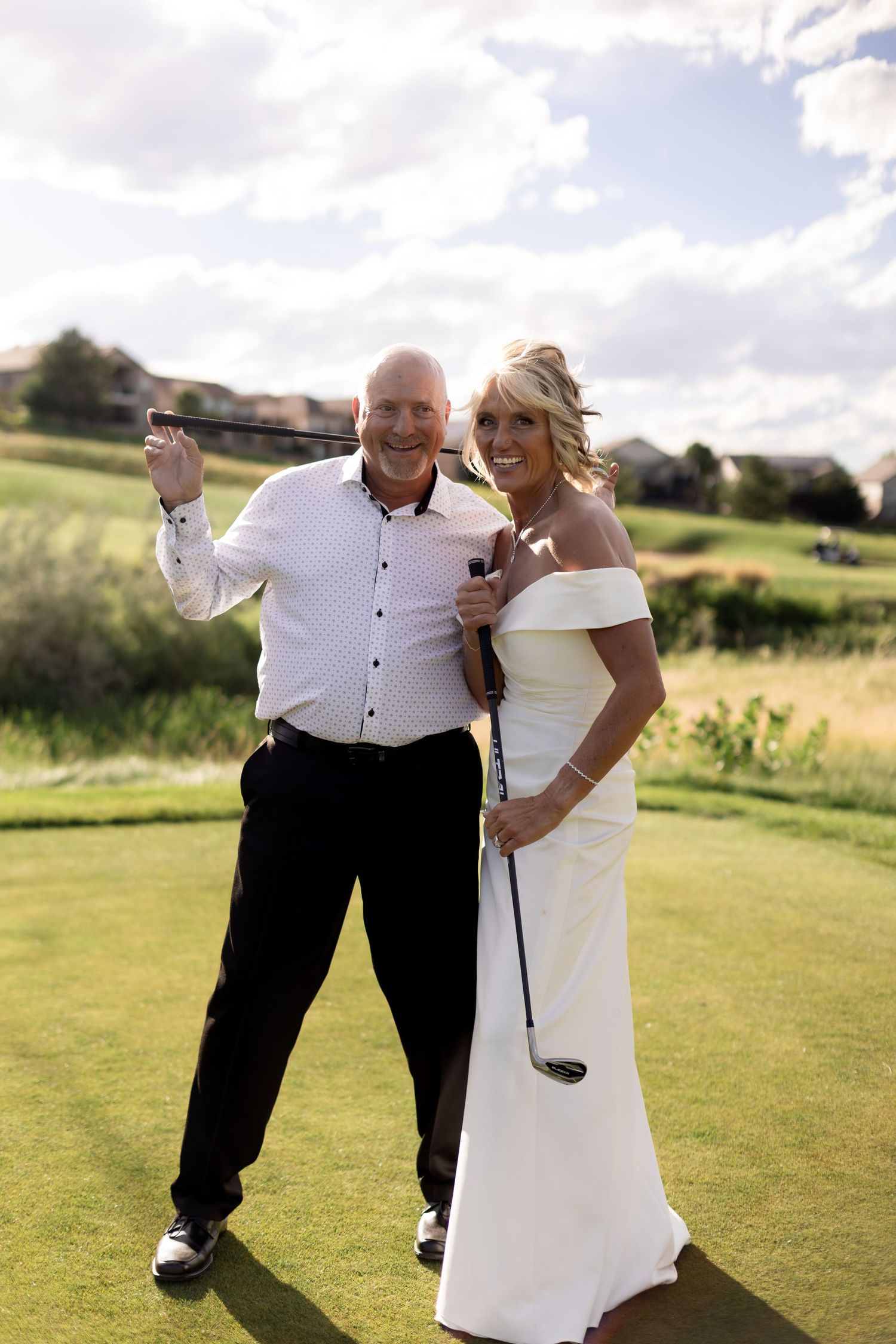 A smiling couple in wedding attire poses with golf clubs on a sunny golf course, bride in white gown, groom in black pants.