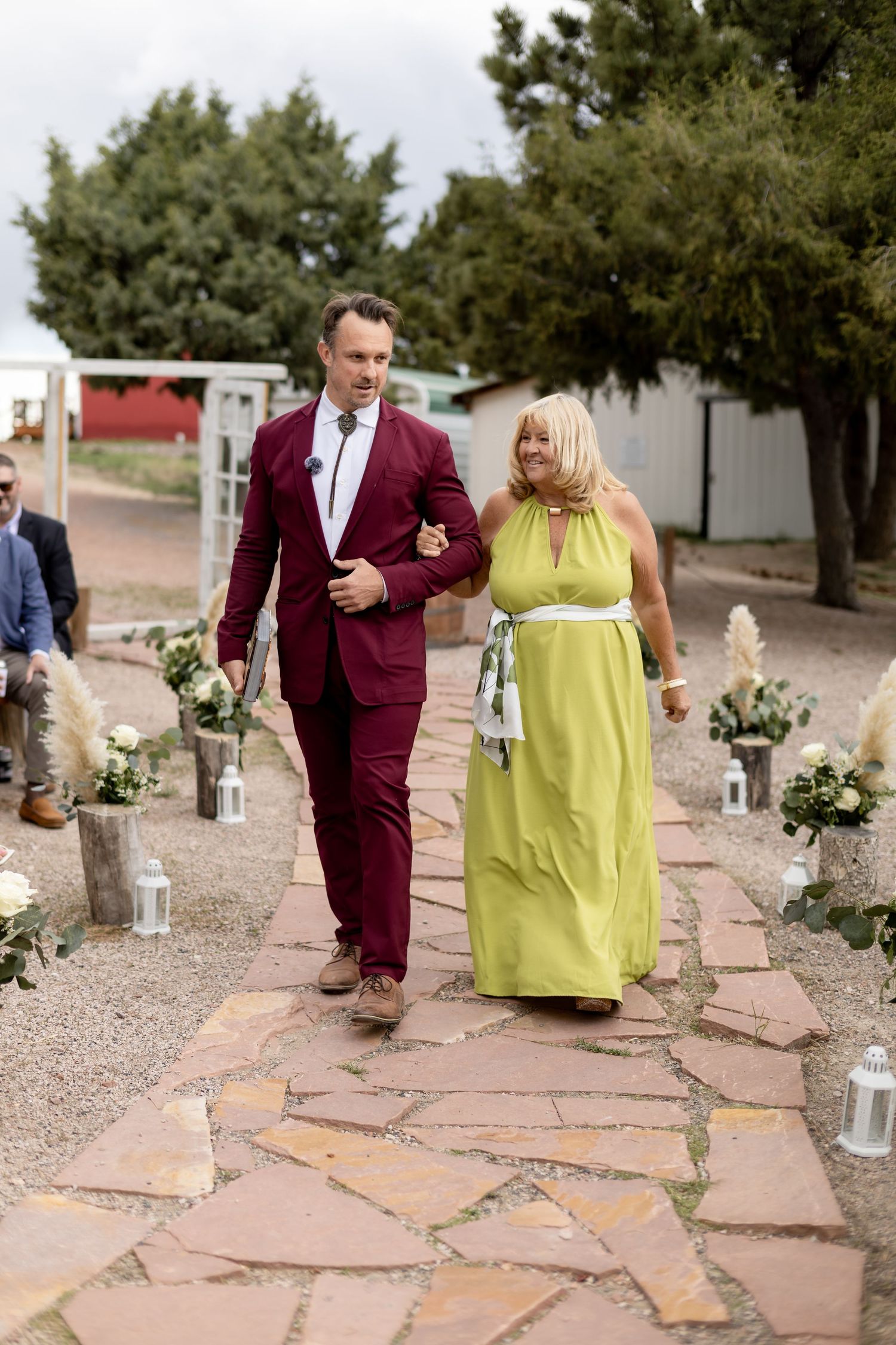Groom in burgundy suit escorts woman in yellow-green gown down stone aisle at outdoor wedding.