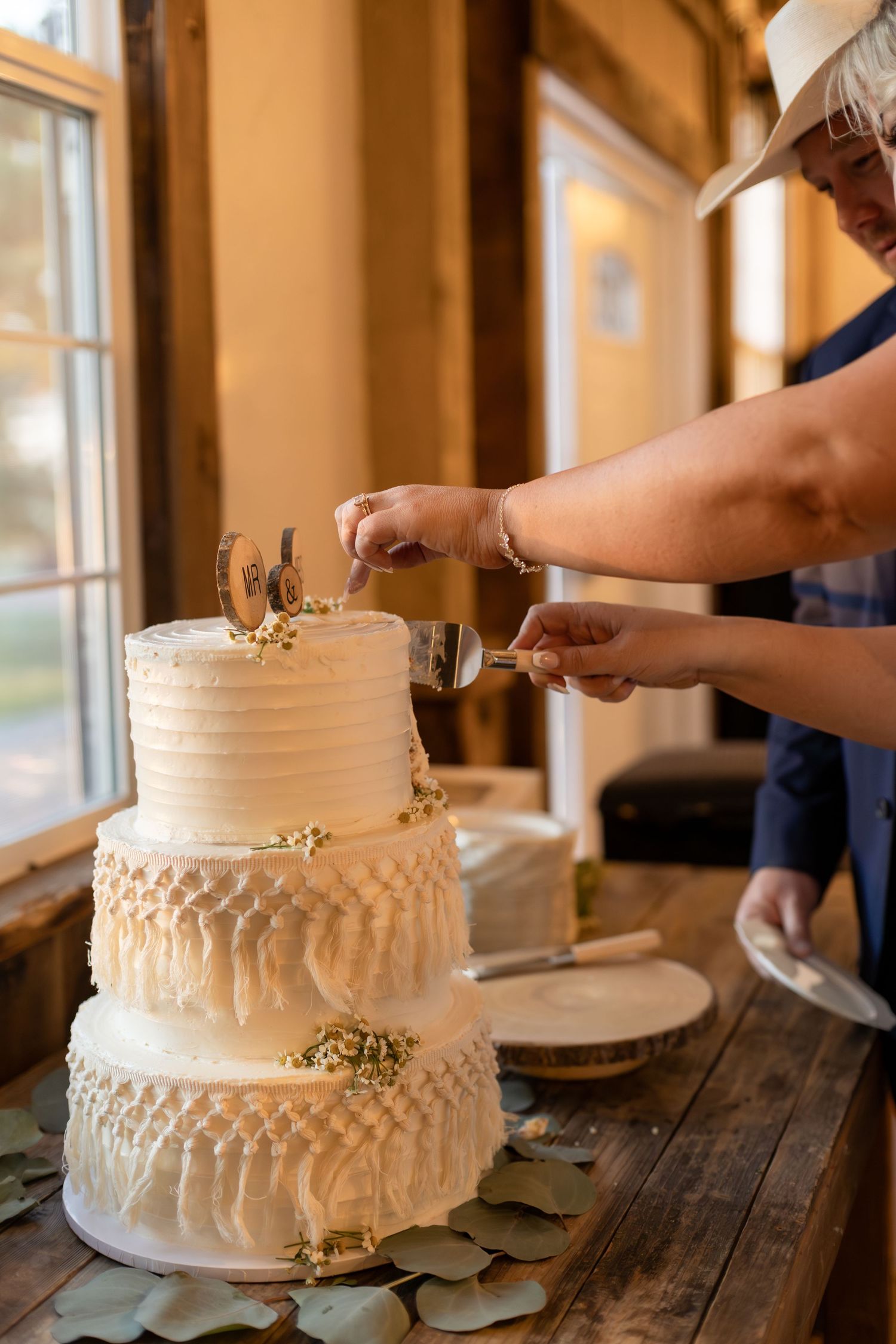 Bride and groom cutting elegant three-tier white wedding cake with macrame details and horseshoe topper.