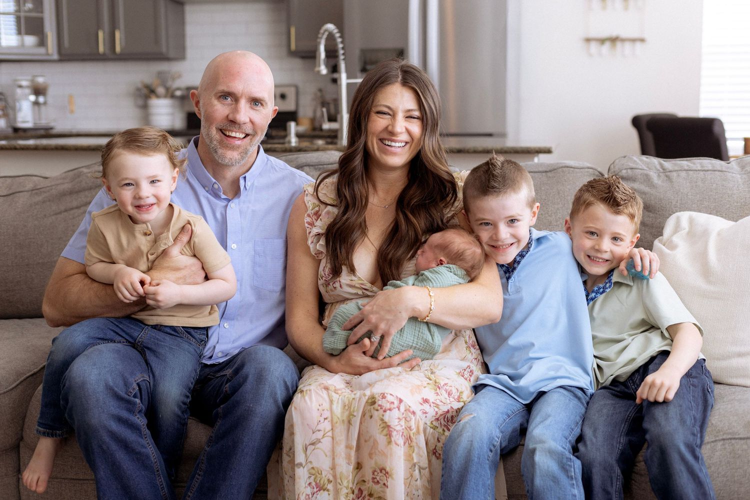 A family sits together on a gray couch in their living room with a modern kitchen visible in the background.