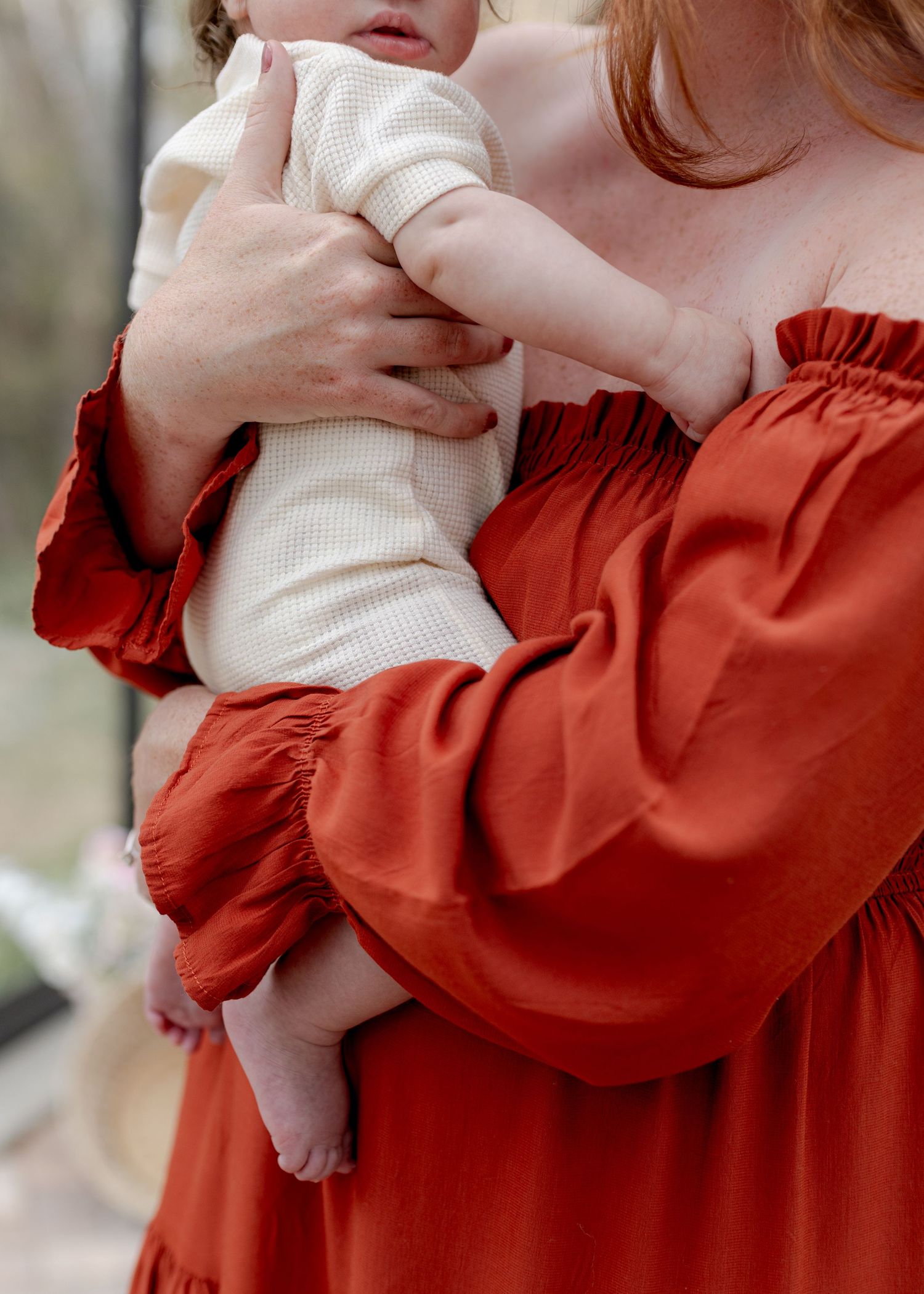 A woman in a rust red off-shoulder dress tenderly holds a baby dressed in a cream waffle knit outfit.