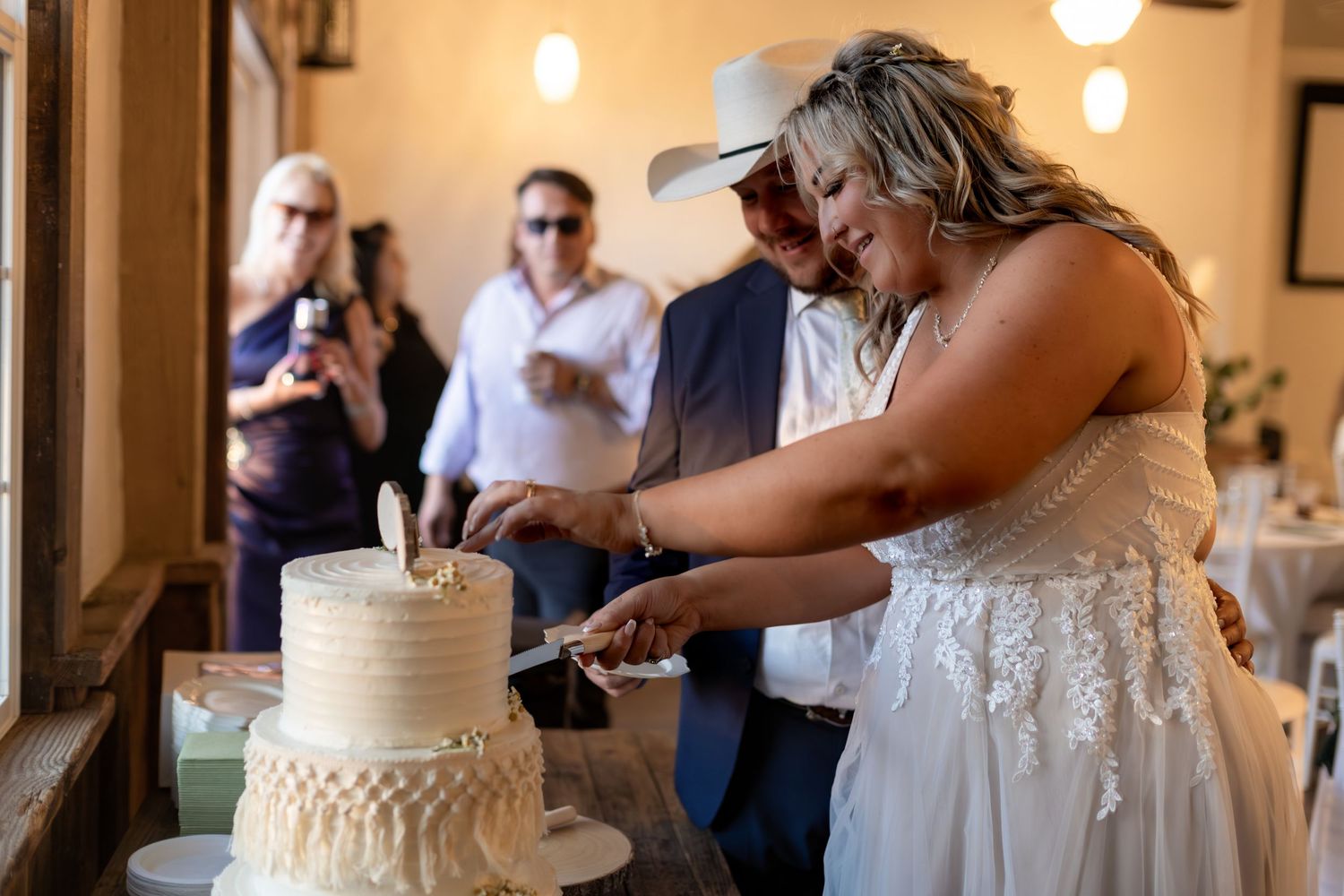 Bride and groom cutting their white wedding cake together while guests watch and photograph.