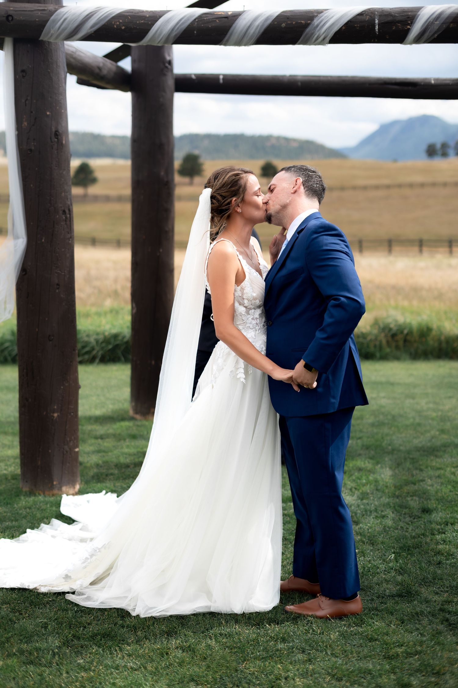 Bride and groom share first kiss under rustic wooden pergola with scenic mountain meadow backdrop.