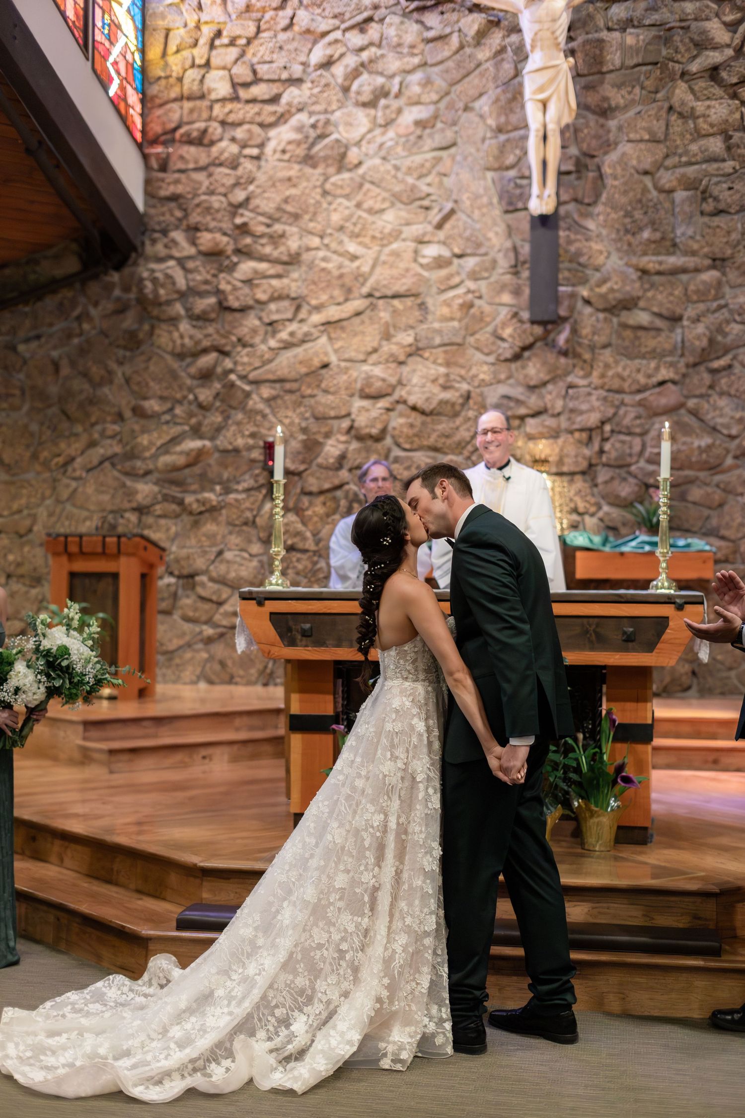 Bride and groom share first kiss at church altar during wedding ceremony with officiant smiling behind them.