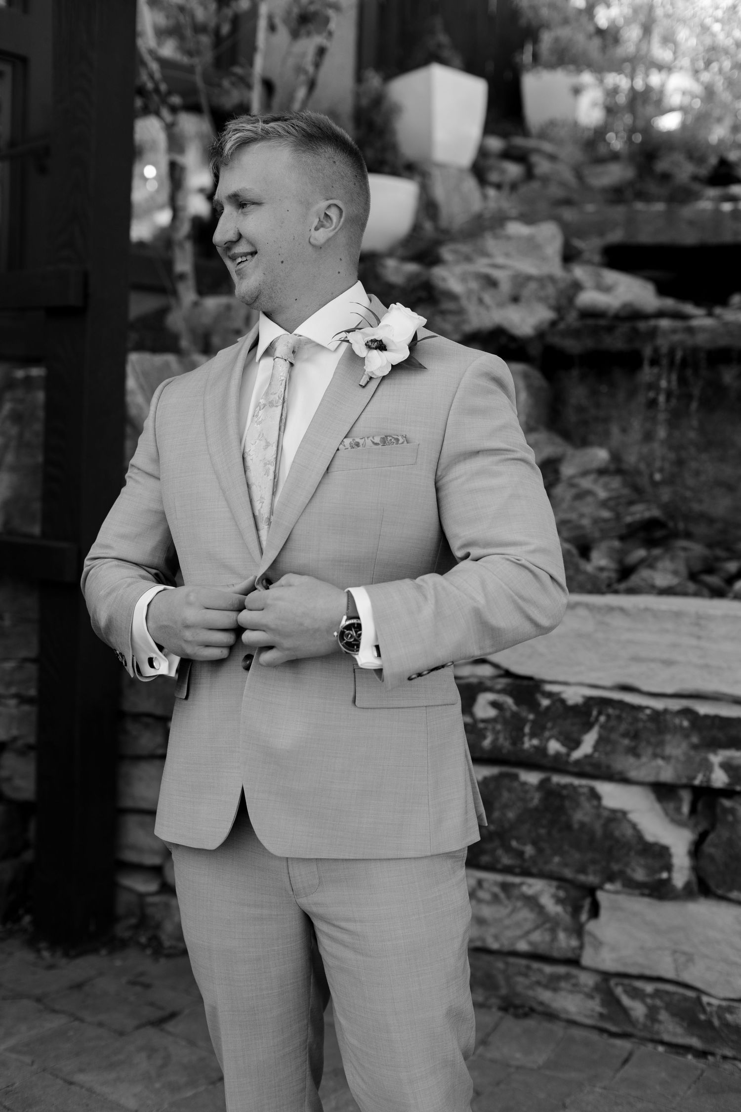 Groom in tan suit with floral boutonniere adjusting jacket in black and white outdoor portrait.