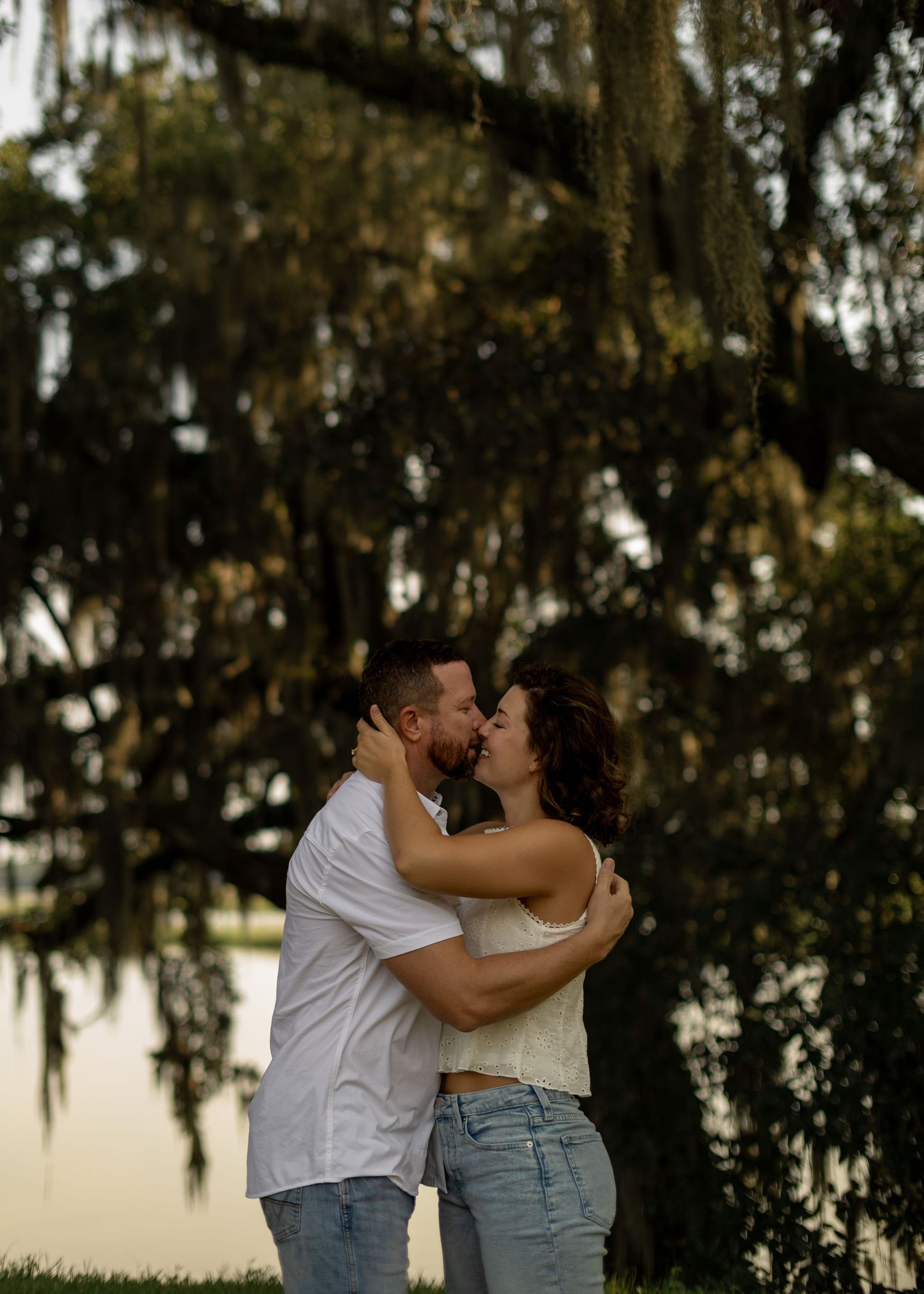 Couple embracing and kissing under Spanish moss-draped oak trees near a lake during golden hour.