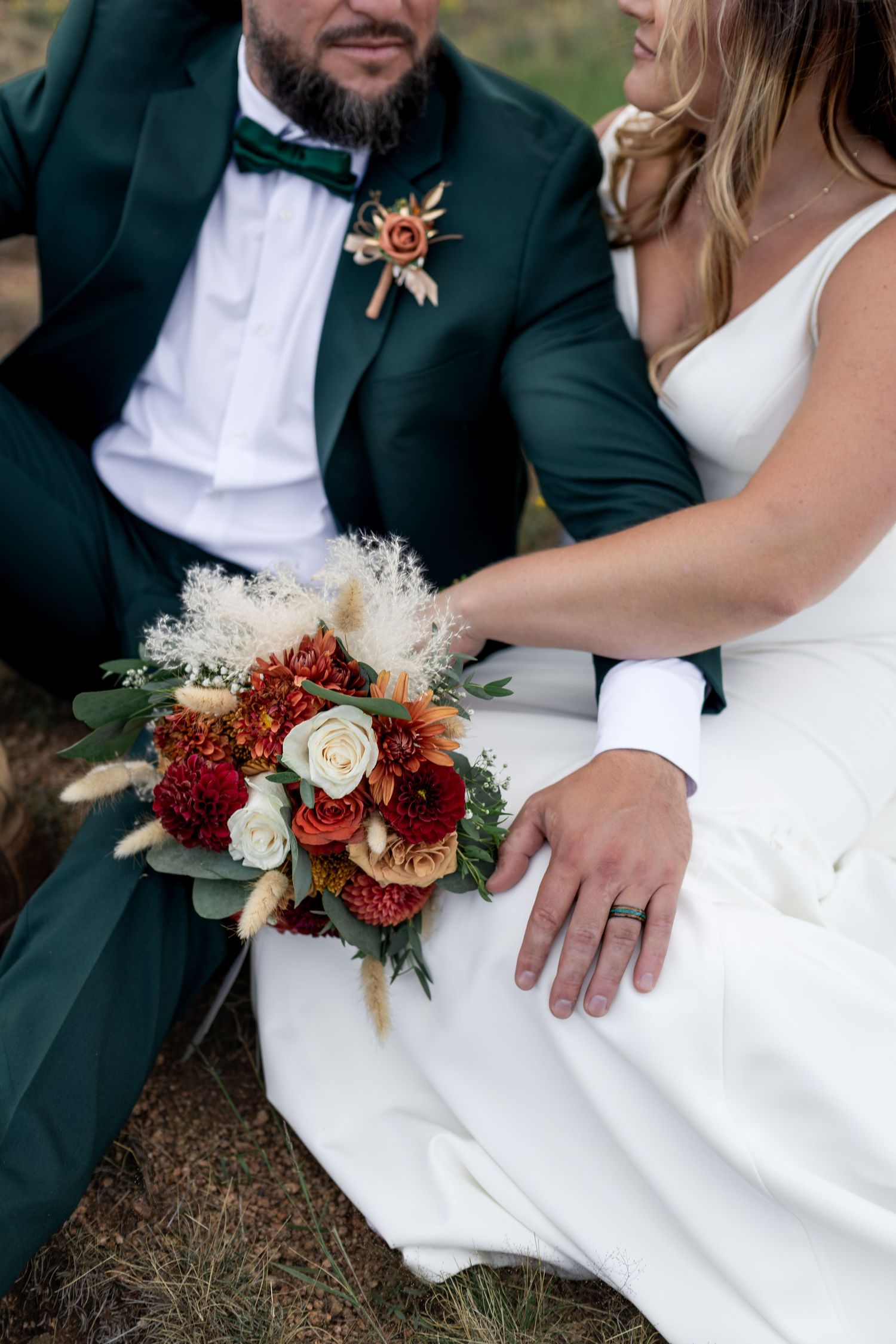 Bride and groom sitting together holding a rustic bouquet of red, orange, and white roses with pampas grass accents.