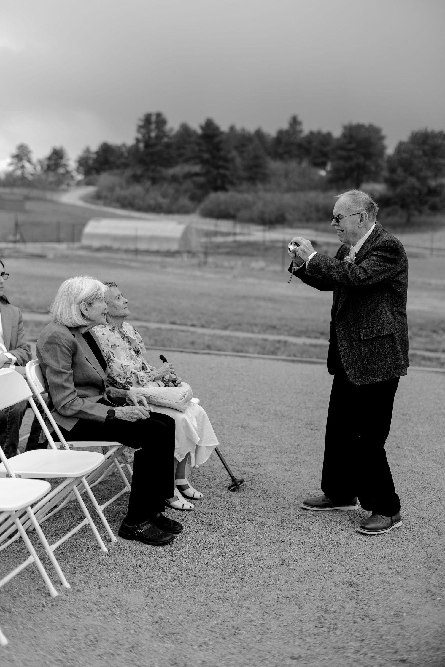 Elderly man photographing two women seated in folding chairs at an outdoor event, black and white photo.