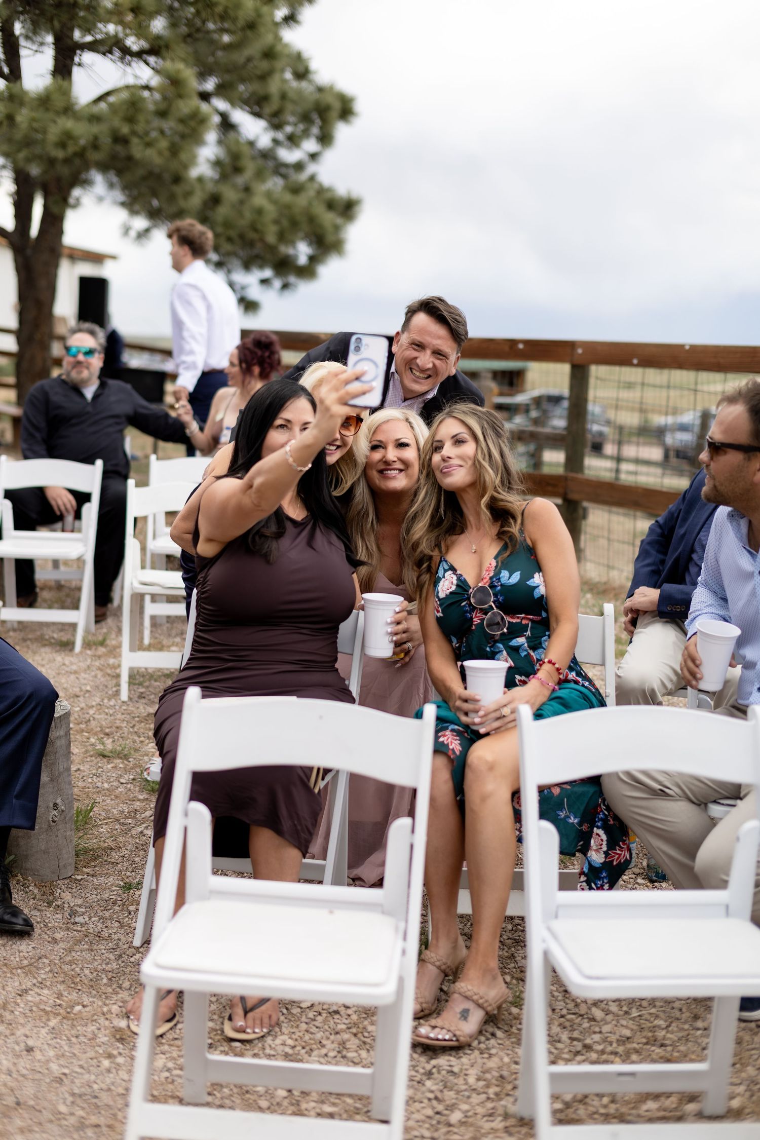 Wedding guests take a group selfie with a smartphone while seated in white folding chairs at an outdoor ceremony.