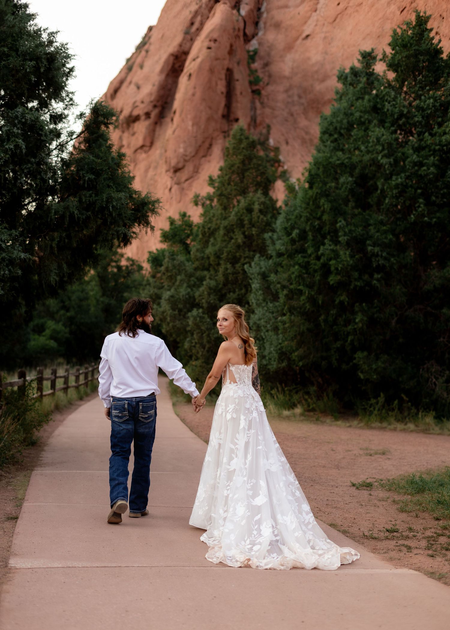 Bride in lace gown and groom in jeans hold hands walking a path at Garden of the Gods, Colorado.