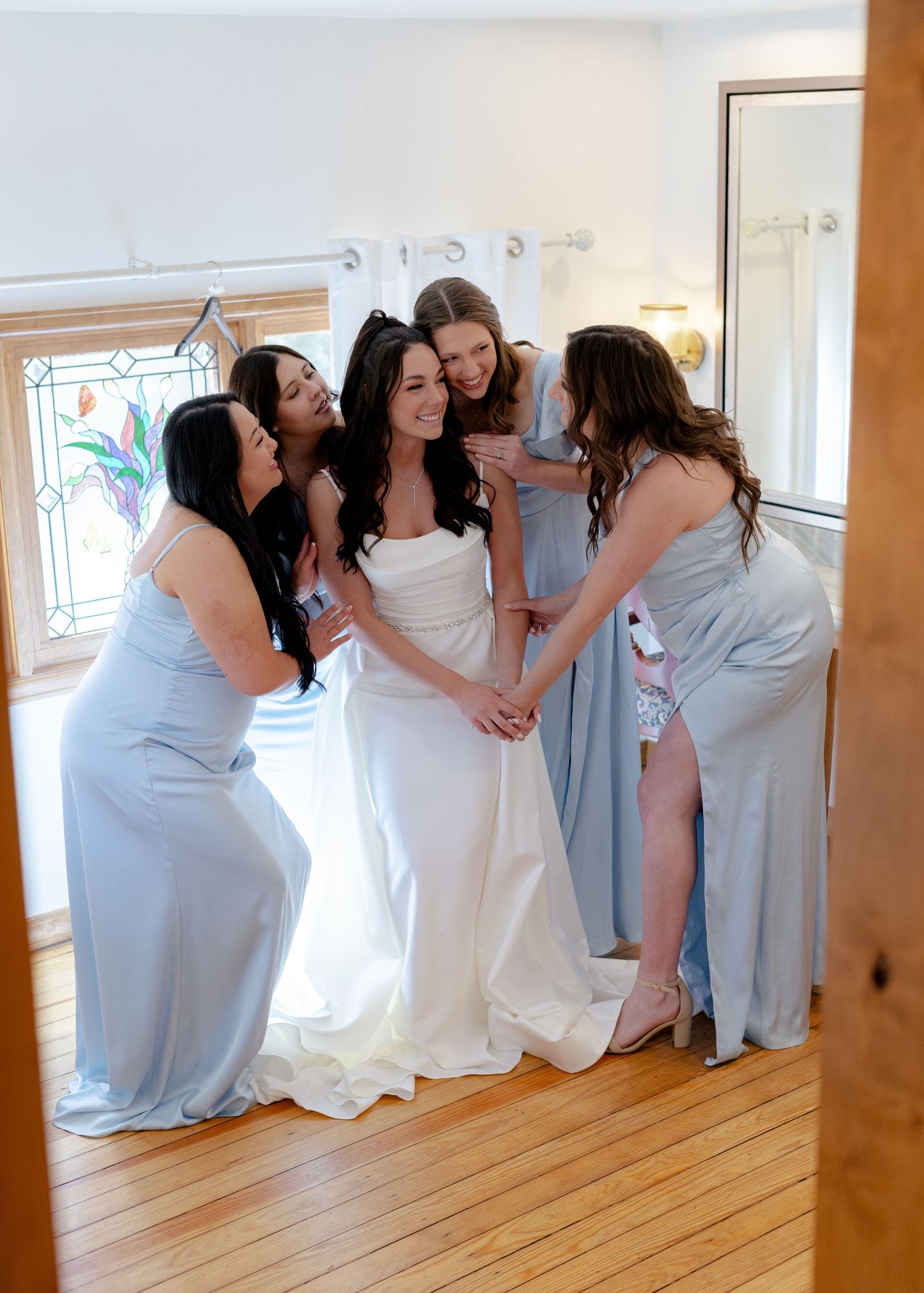 Bride in white gown surrounded by bridesmaids in light blue dresses sharing a joyful moment indoors.