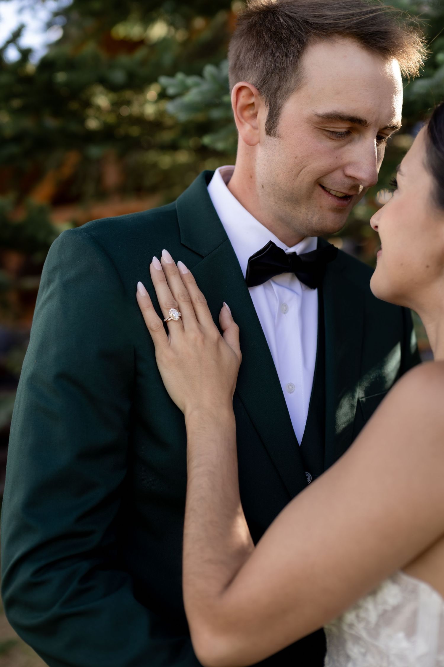 Bride places hand on grooms green tuxedo jacket showing diamond engagement ring outdoors.