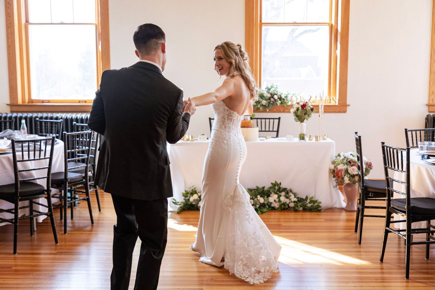 Bride and groom share their first dance in a sunlit wedding reception hall with floral decorations.