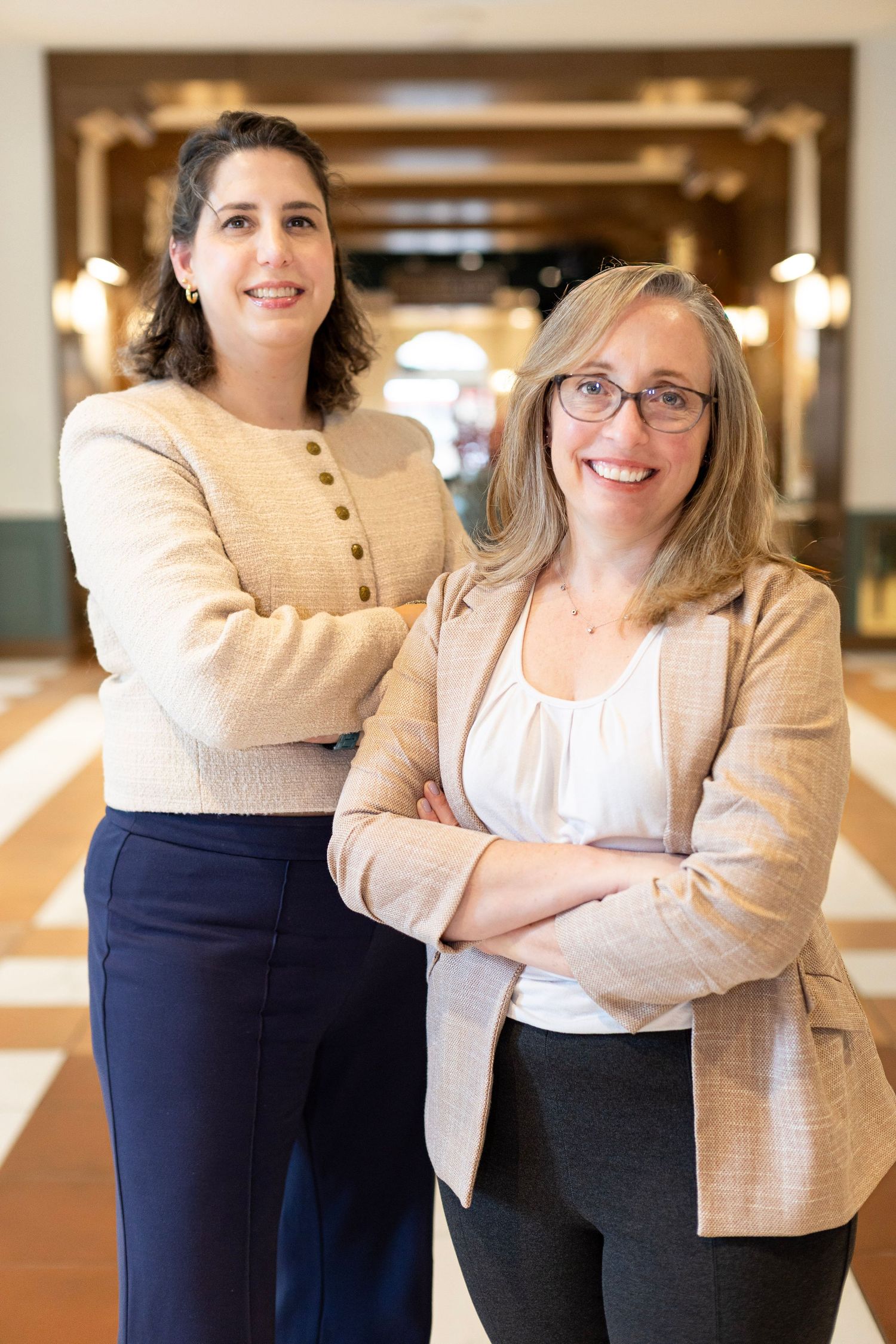 Two professional colleagues in business attire pose confidently in an office hallway.