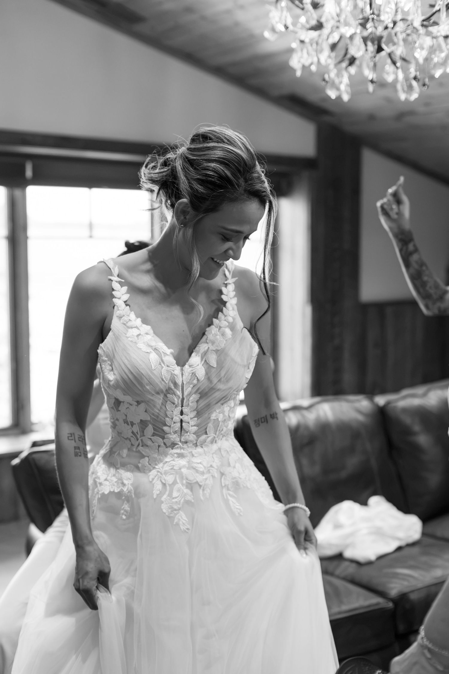 Bride in lace wedding gown smiling down in black and white photo inside rustic venue with chandelier.