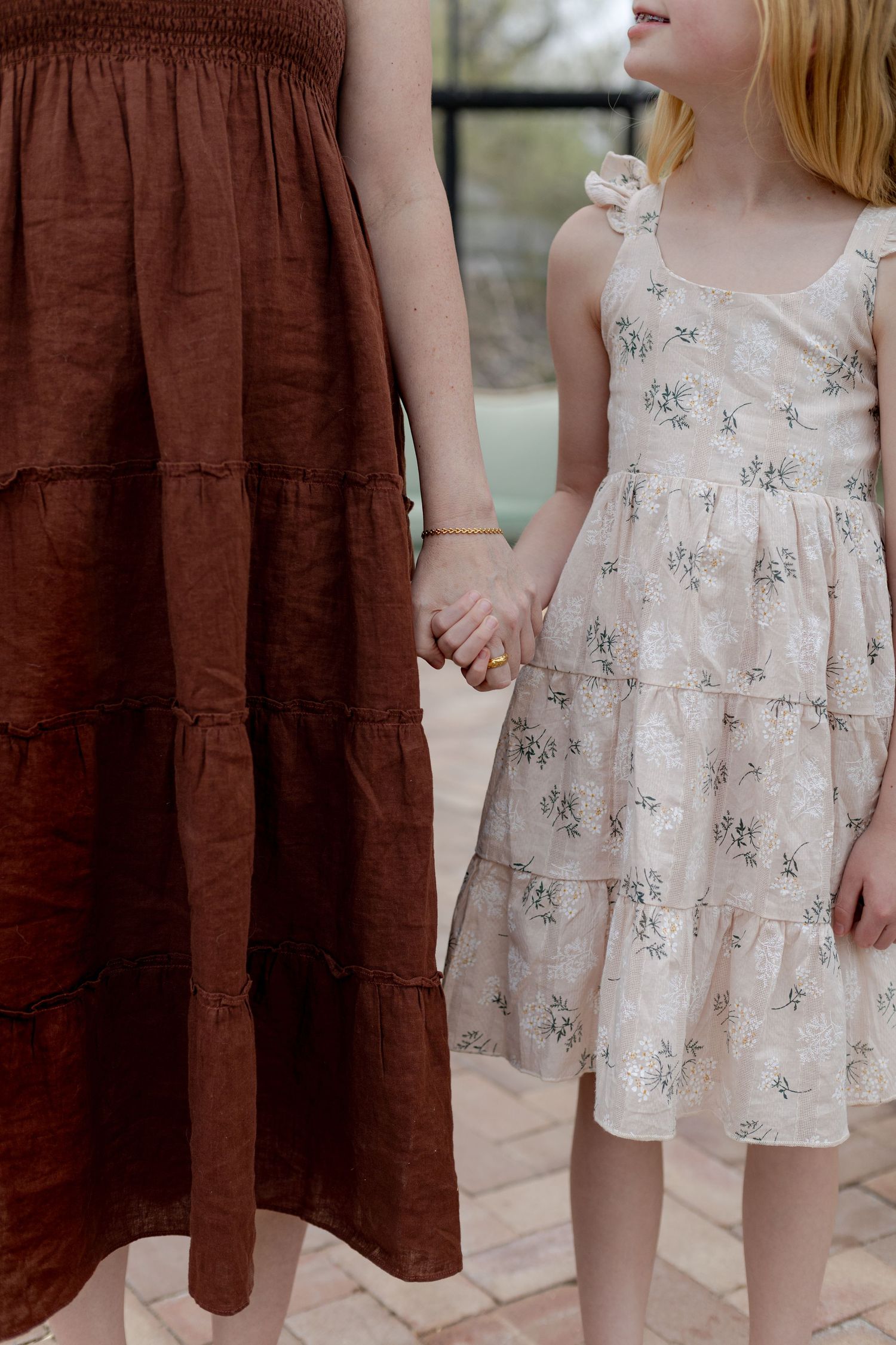 A woman in a brown tiered maxi dress holds hands with a young girl in a floral cream dress.