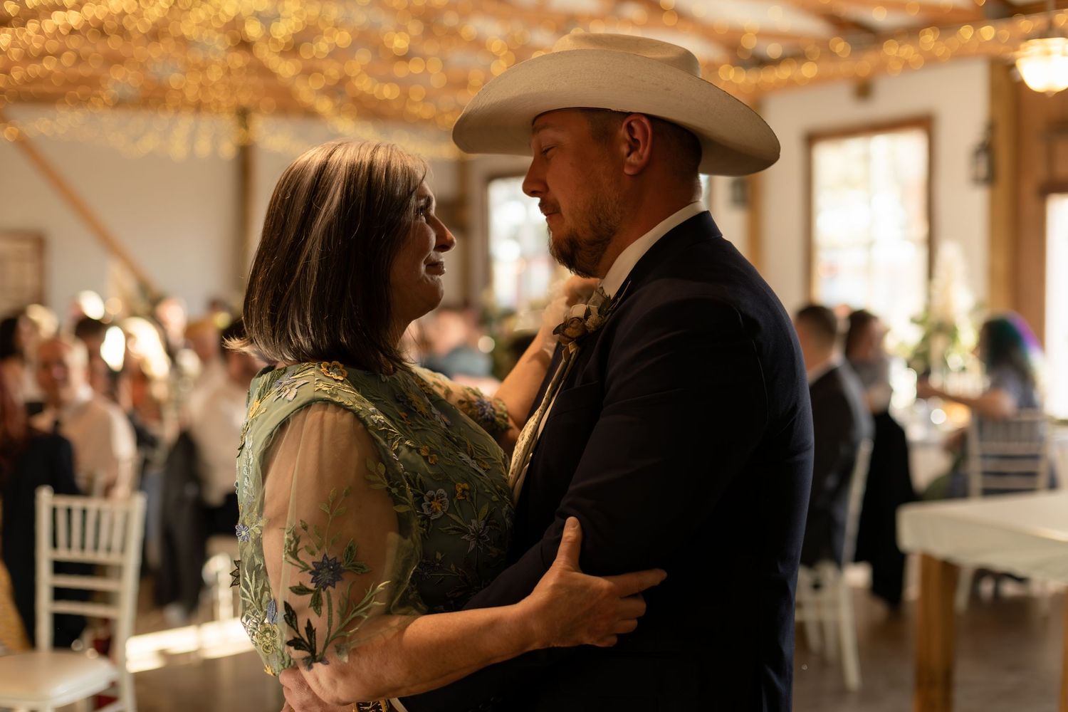 Groom in white cowboy hat shares romantic dance with partner under warm string lights at wedding reception.