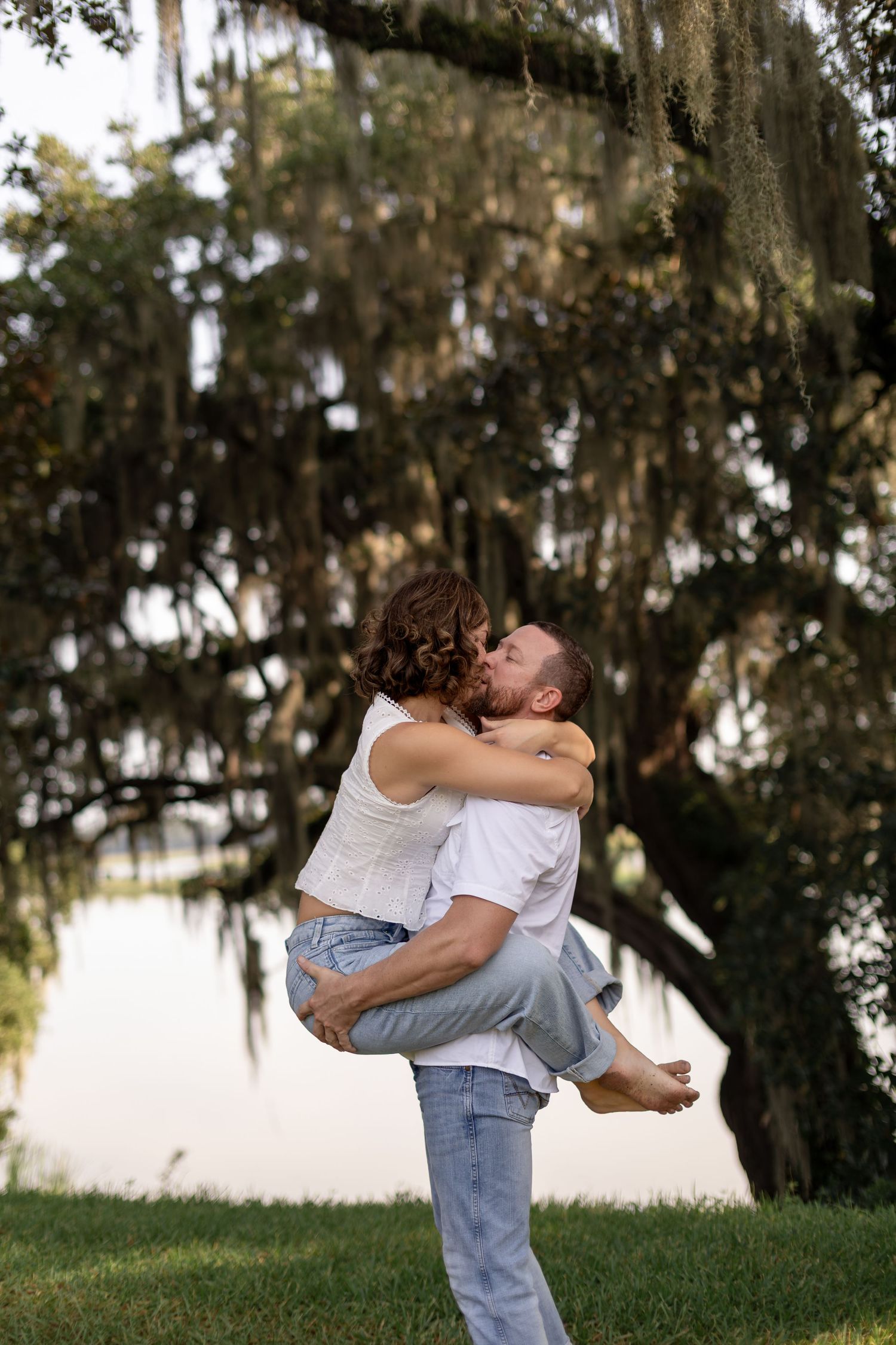 Man lifts woman for a kiss under Spanish moss-draped oak trees near a serene pond outdoors.