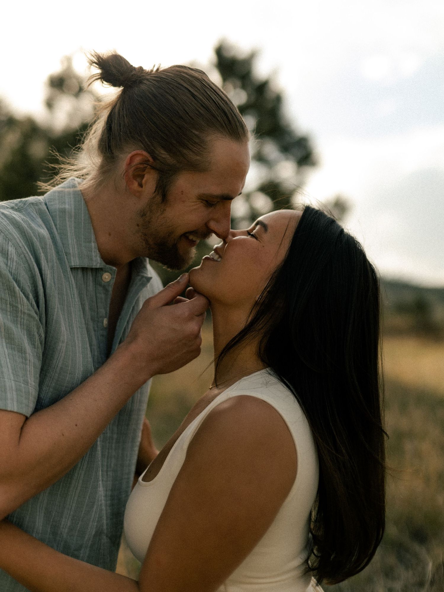 Couple sharing a romantic almost-kiss moment in a golden field at dusk, man with bun holding woman's chin.