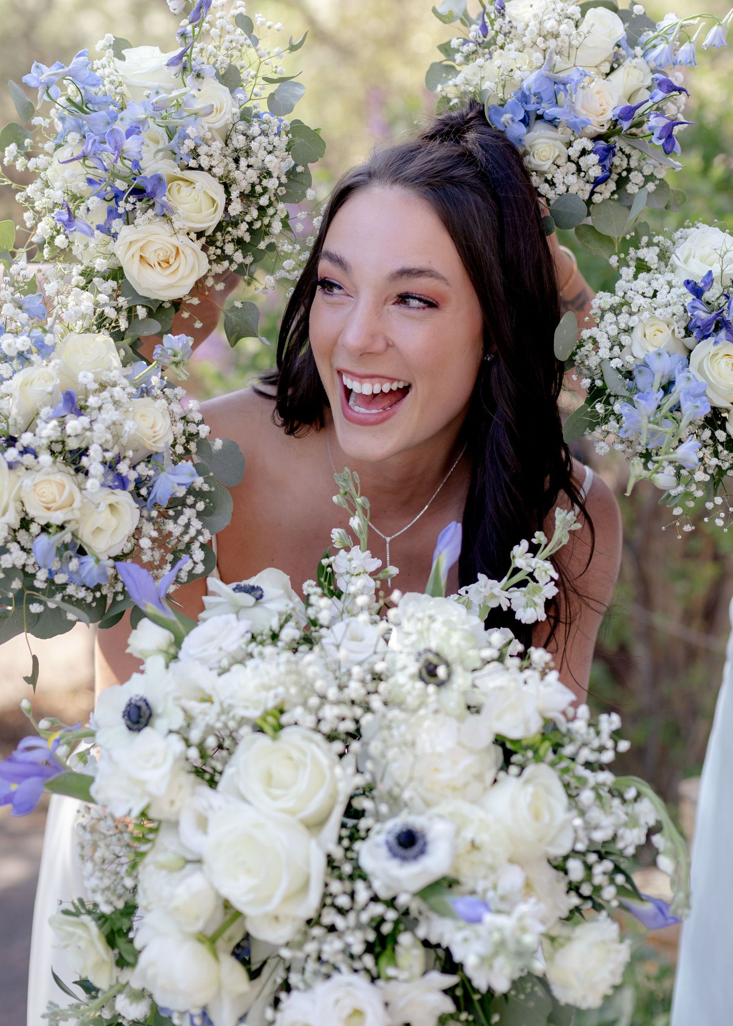 Smiling bride surrounded by white and blue floral bouquets with roses and anemones outdoors.