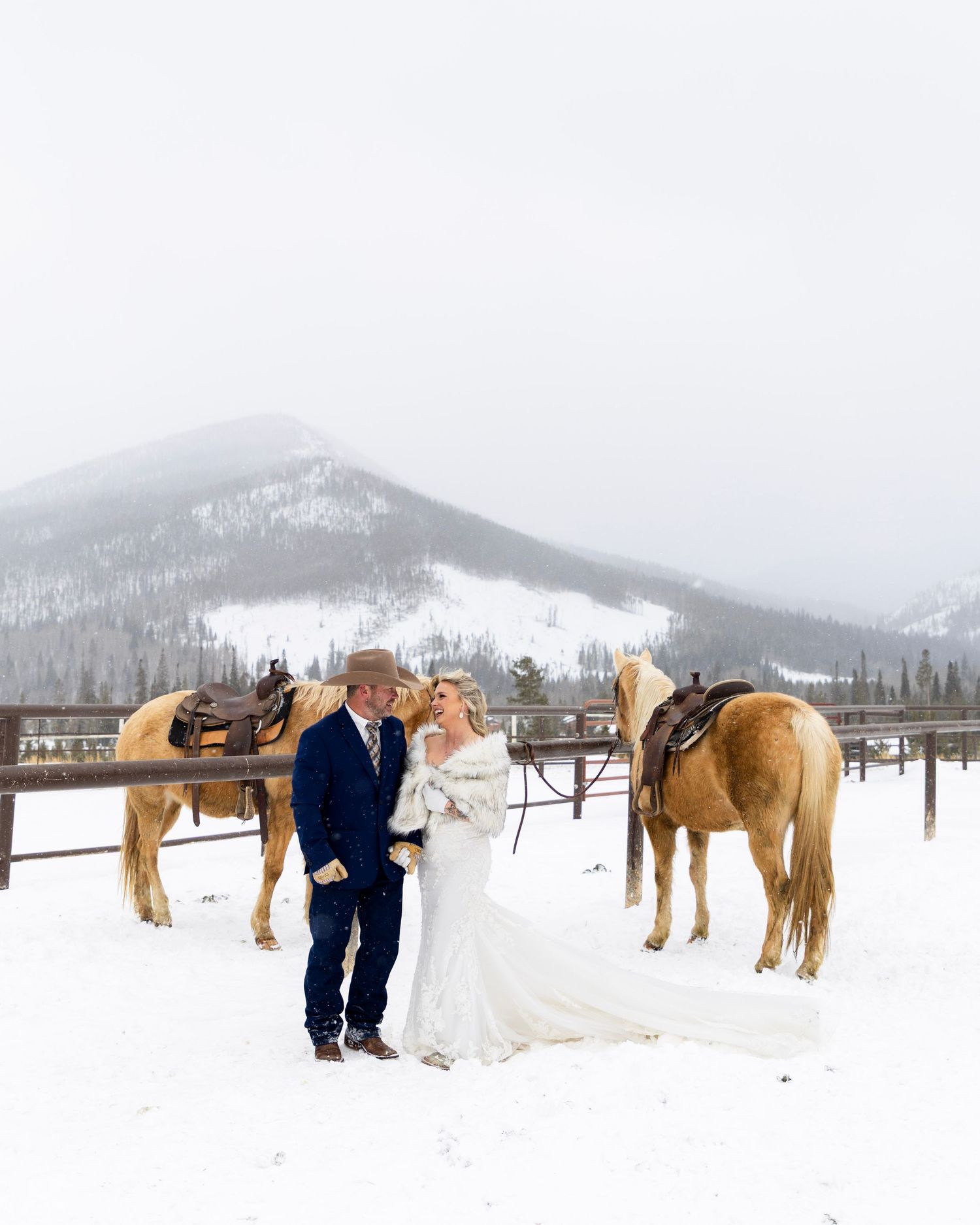 Bride and groom stand with horses in snowy mountain landscape during winter wedding ceremony.