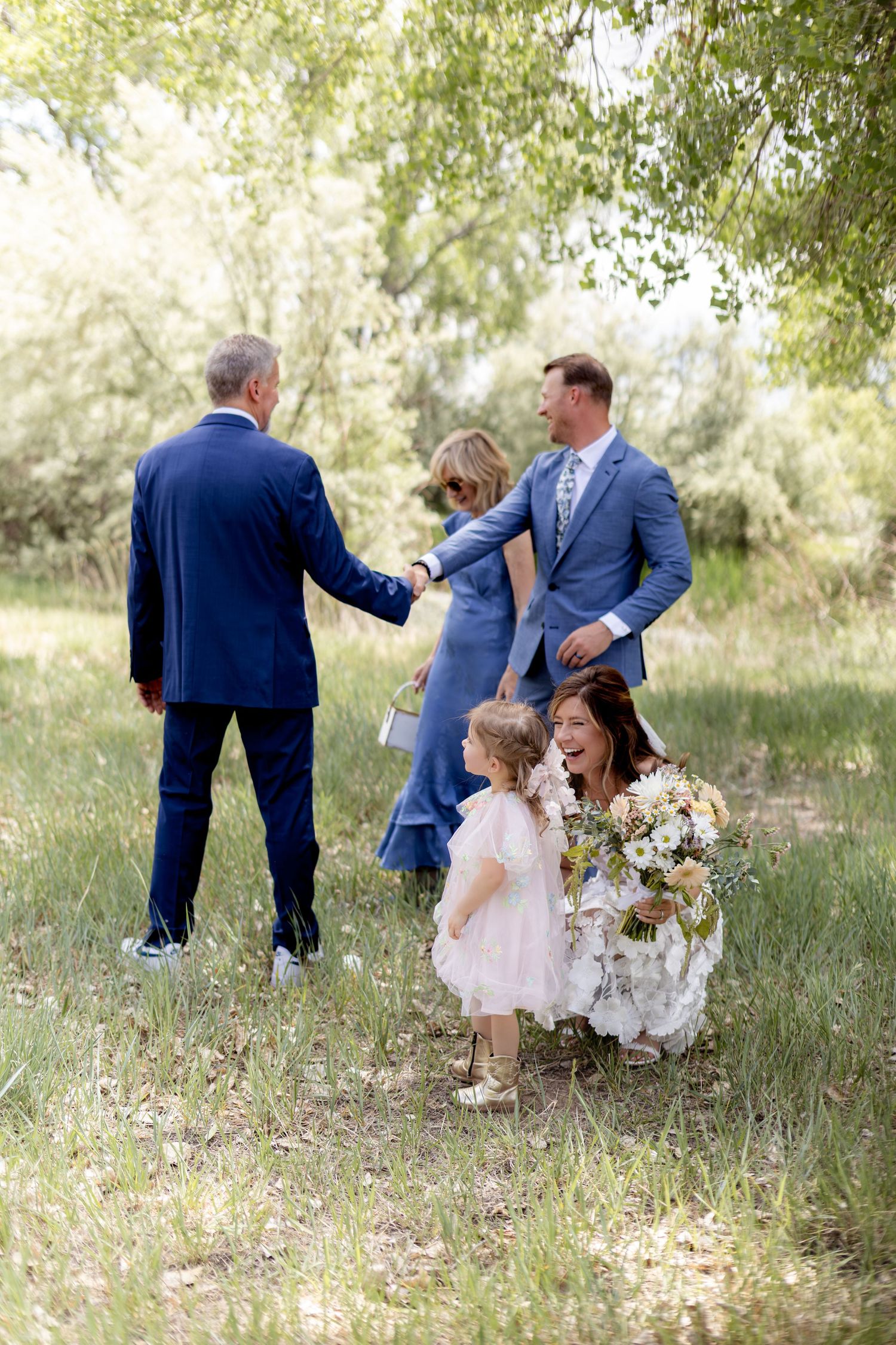 Wedding party in a green park, bride crouches with flower girl while two men in blue suits shake hands.