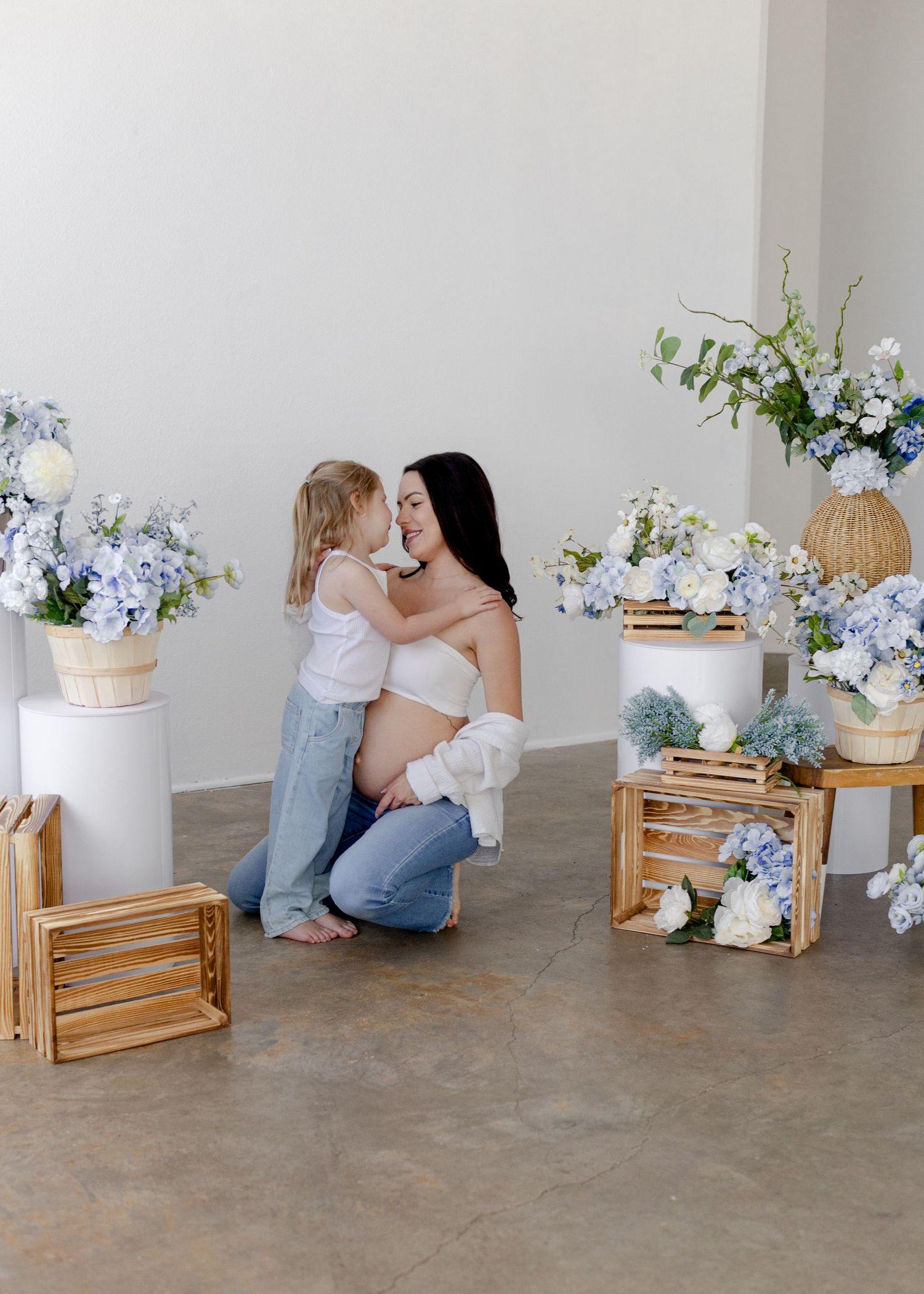 Pregnant woman kneeling with toddler girl among blue hydrangea floral arrangements and wooden crates in studio.