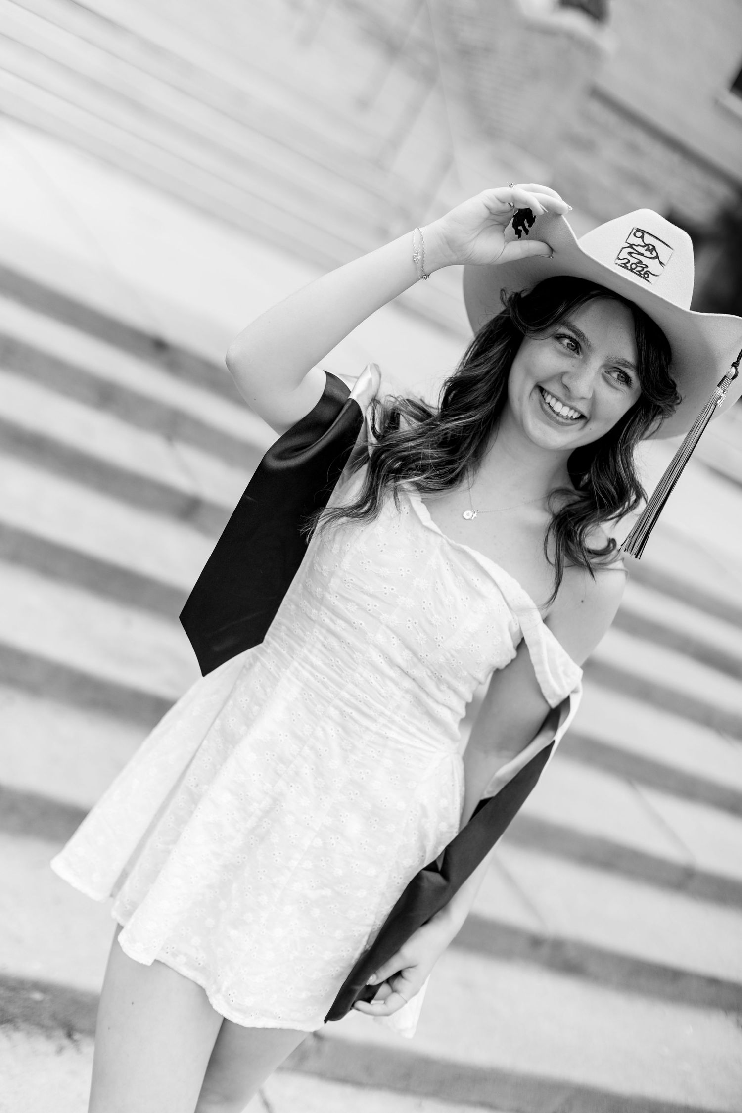 Smiling young woman in white dress and cowboy hat holding graduation gown on marble steps.