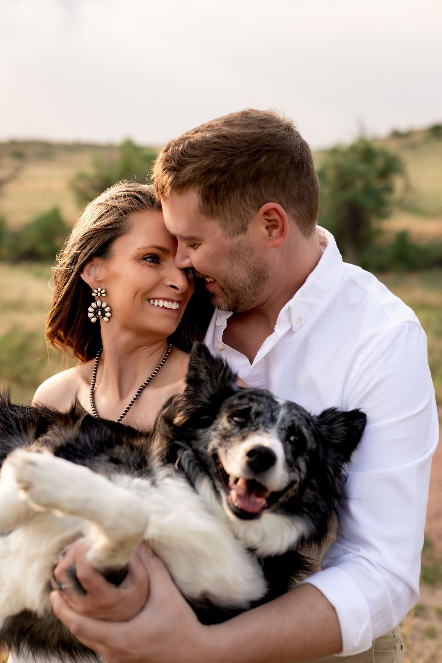 Smiling couple embracing a happy border collie dog outdoors in a scenic field setting.