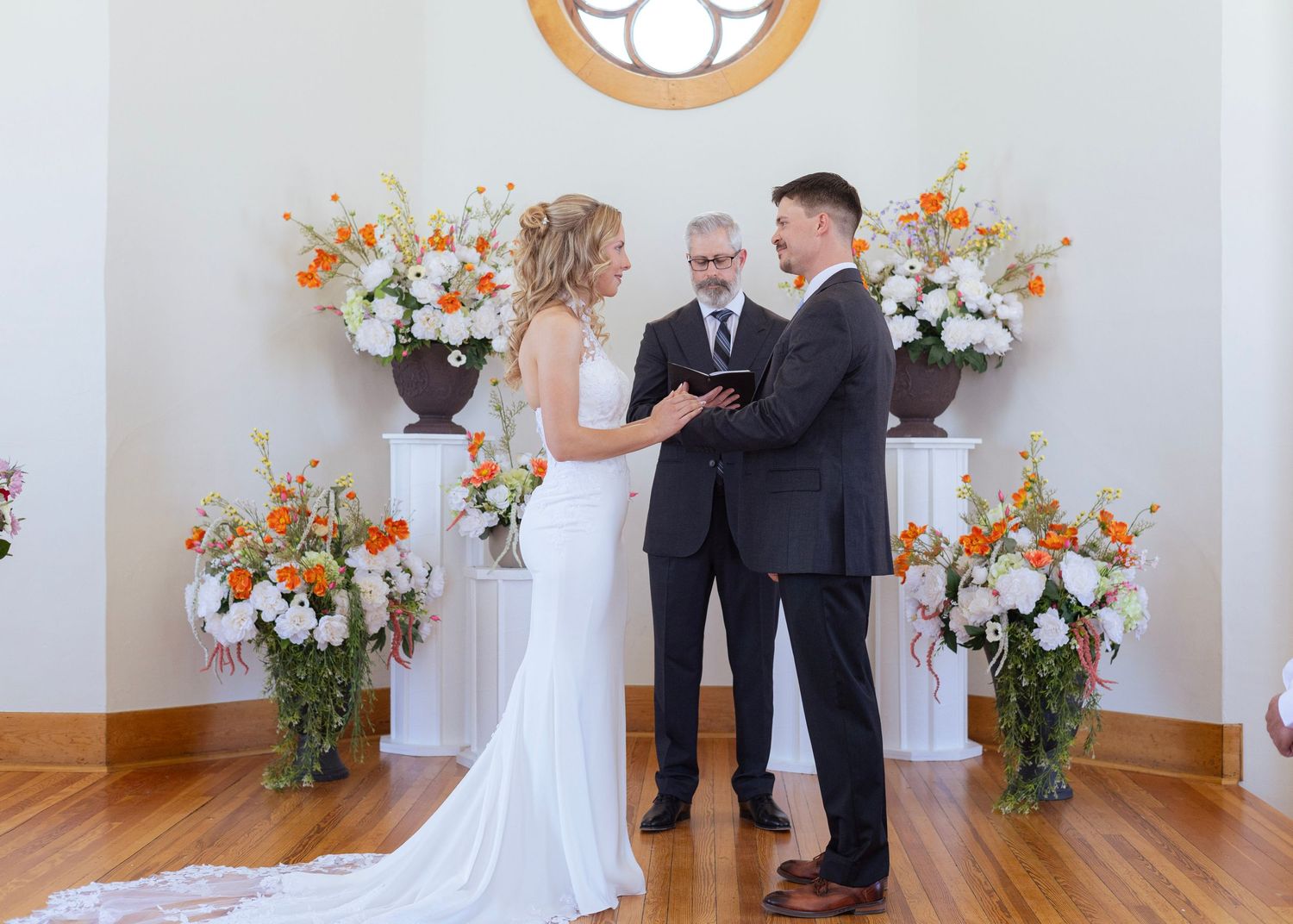 Bride and groom exchange vows at elegant indoor wedding ceremony with orange and white floral arrangements.