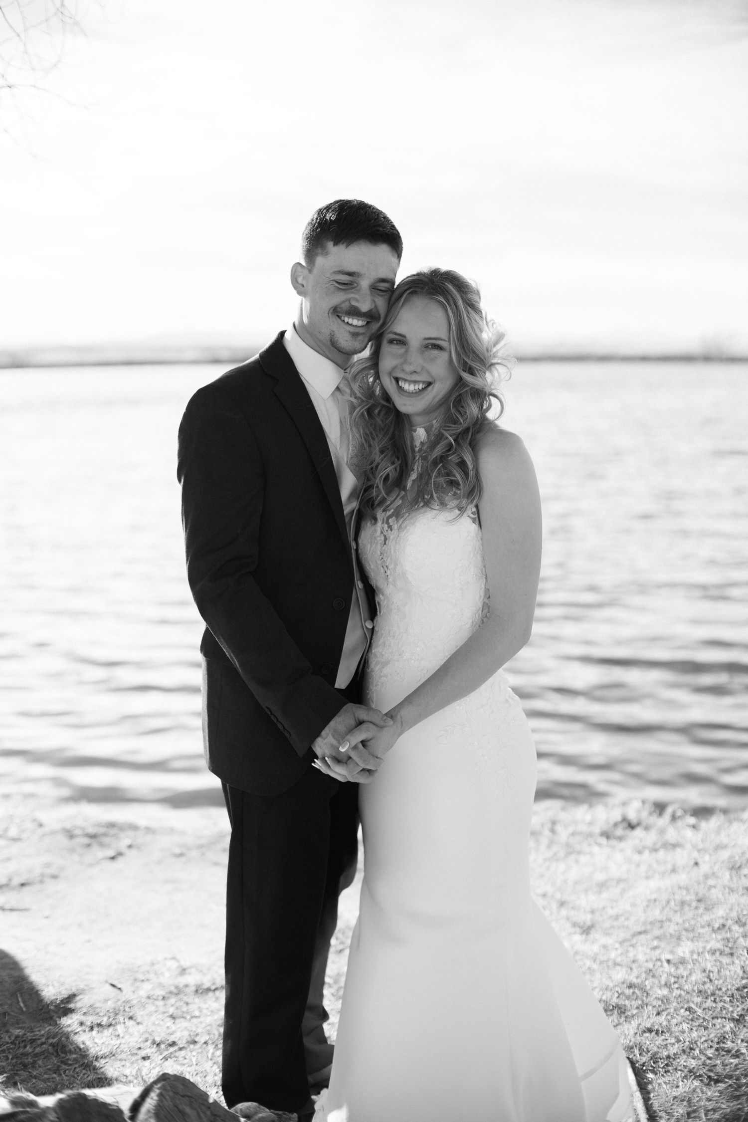 Black and white photo of smiling bride and groom holding hands by a lakeside on their wedding day.