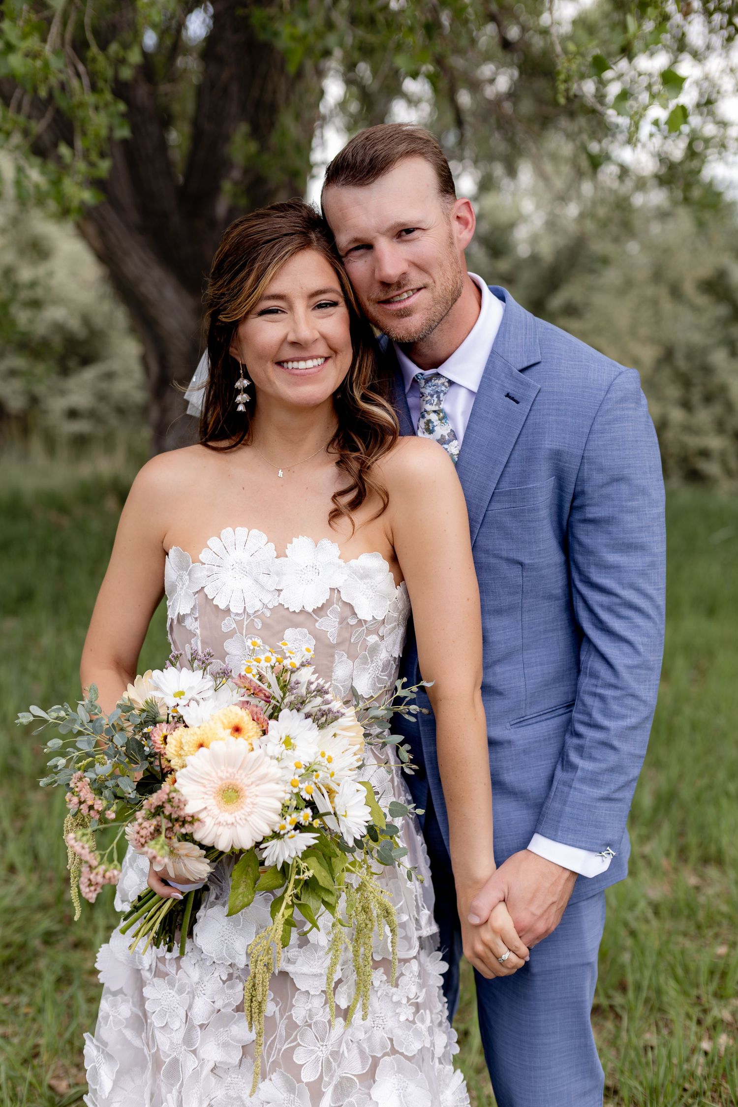 Smiling bride in floral applique gown holds bouquet beside groom in blue suit at outdoor wedding.