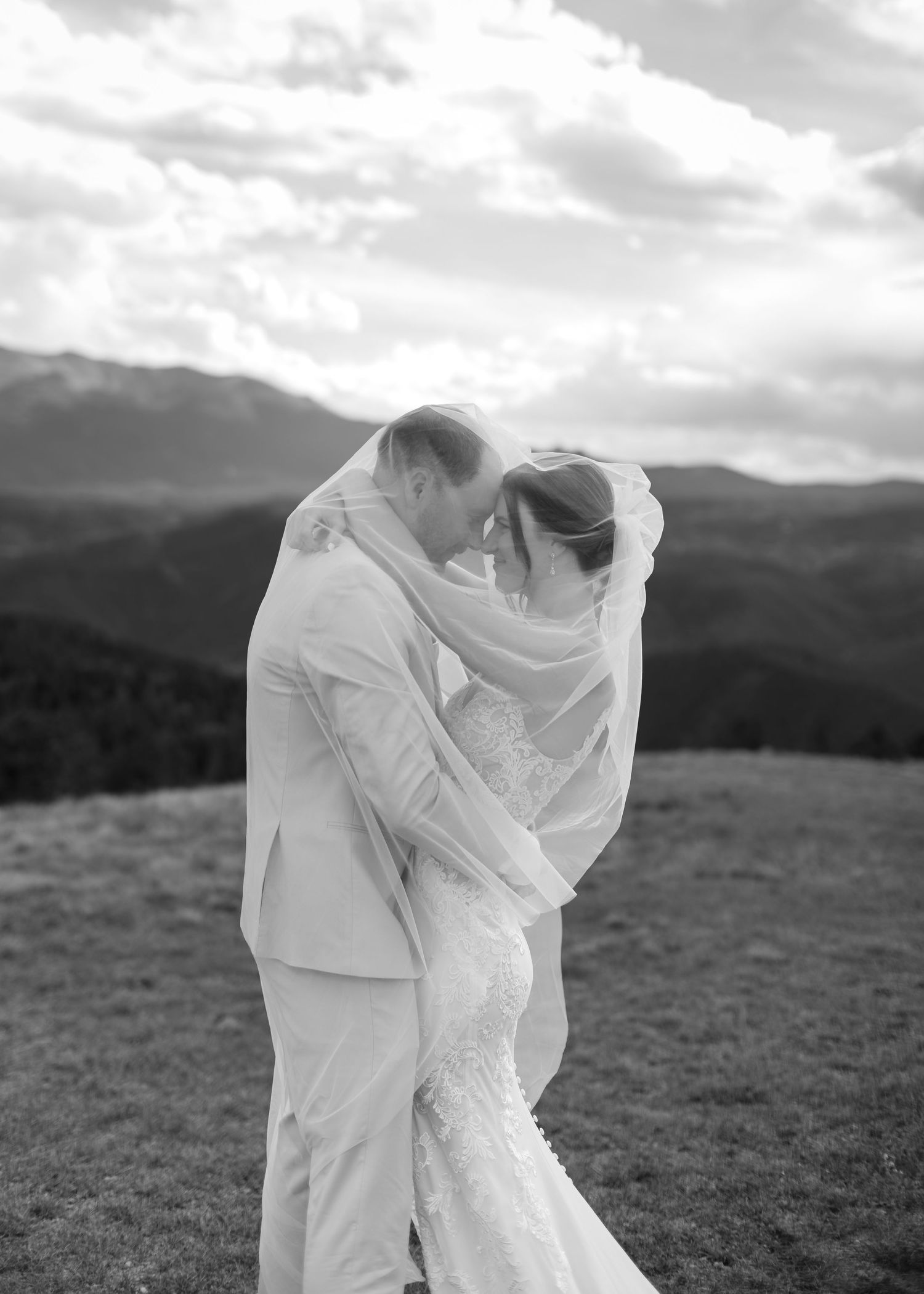 Black and white photo of bride and groom sharing intimate moment under veil with mountain backdrop.
