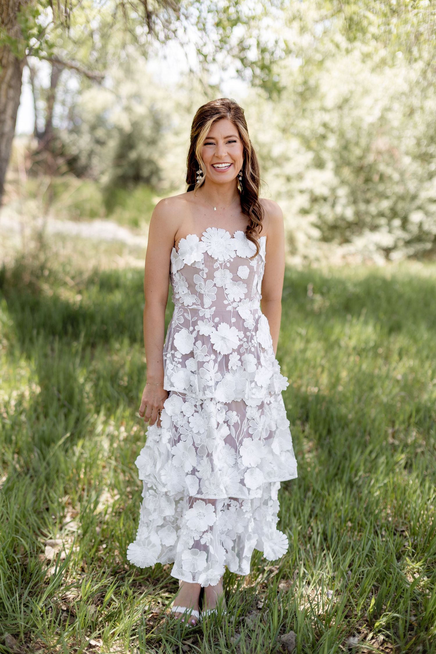 Smiling woman in white 3D floral strapless tiered midi dress standing outdoors in a lush green garden.