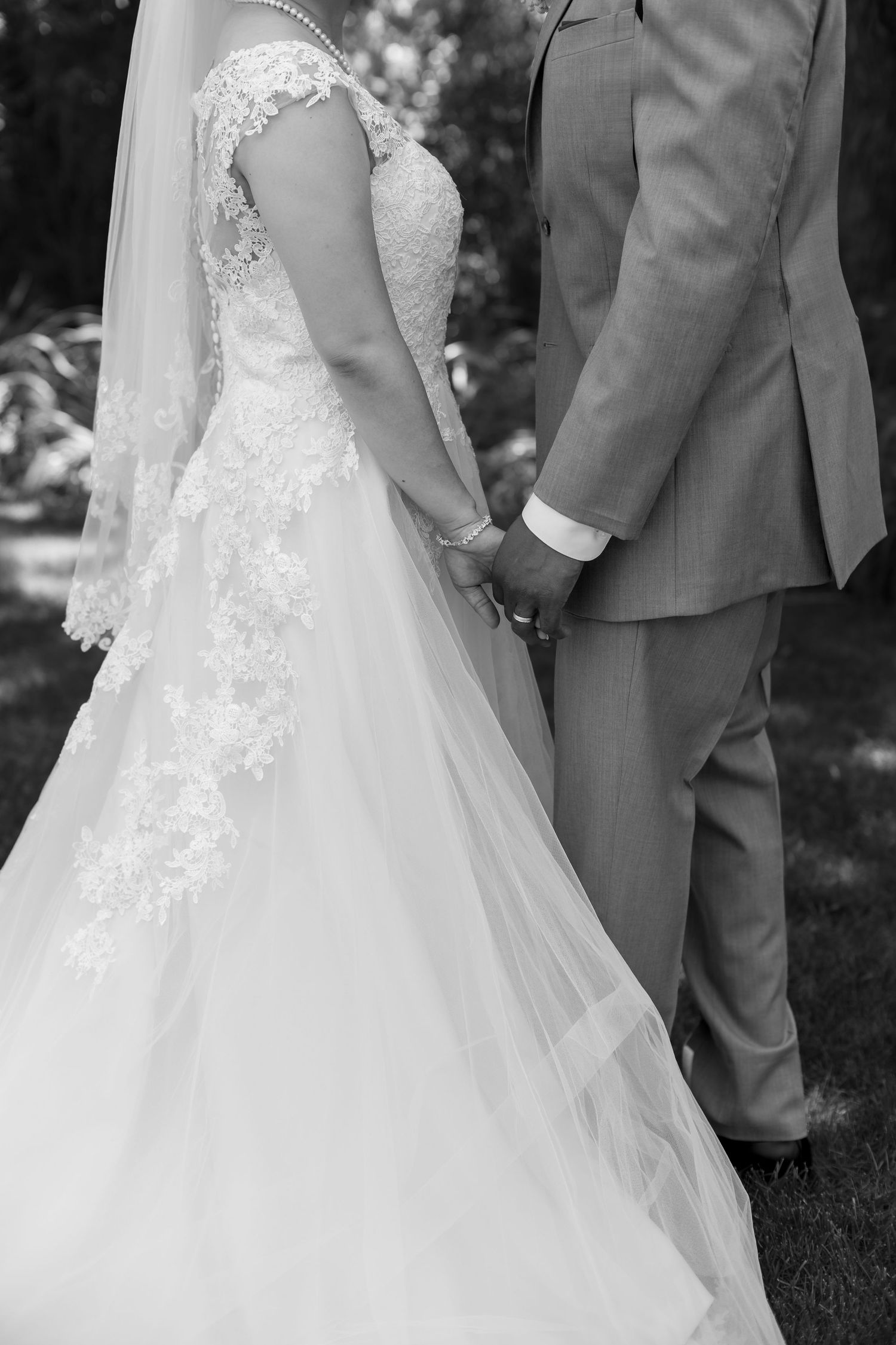 Black and white photo of bride and groom holding hands outdoors, bride wearing lace wedding gown with veil.