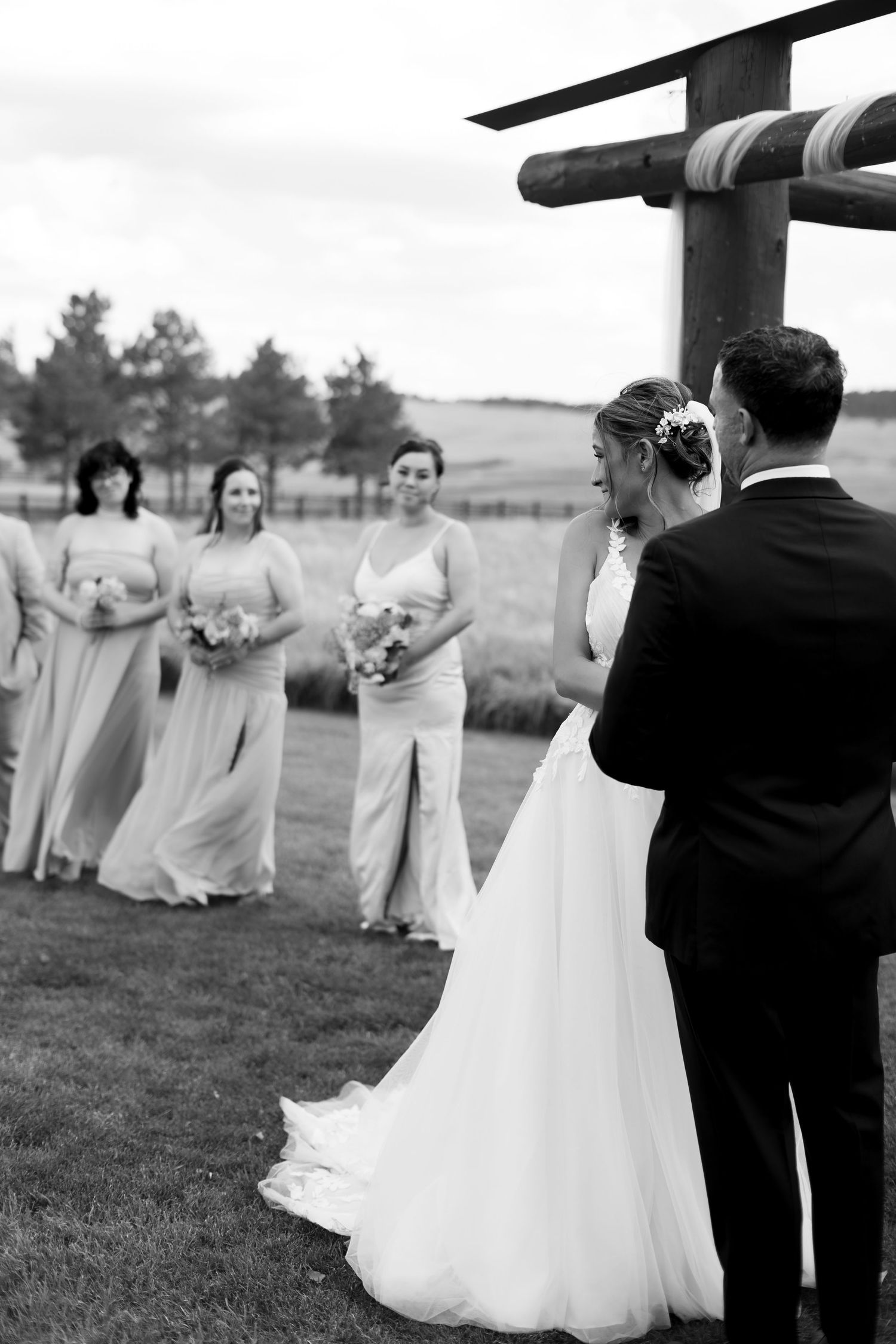 Black and white photo of bride and groom exchanging vows outdoors with bridesmaids in flowing gowns standing nearby.