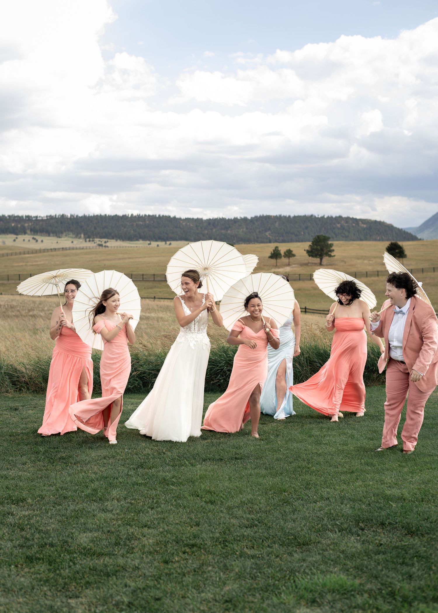Bride and bridesmaids in coral dresses holding white parasols on green lawn with mountain backdrop.