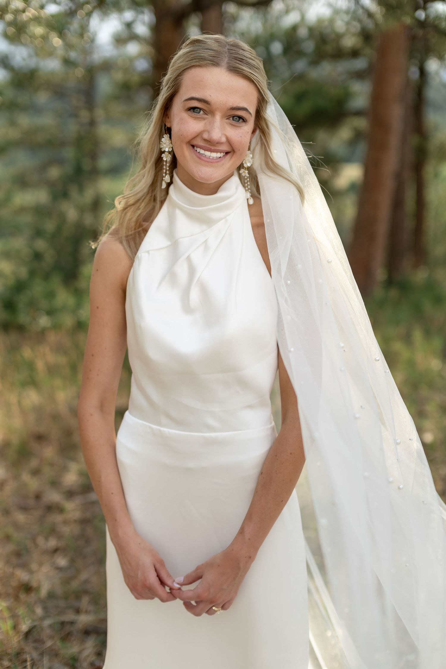 Smiling bride in white satin halter gown and pearl veil poses outdoors among trees.