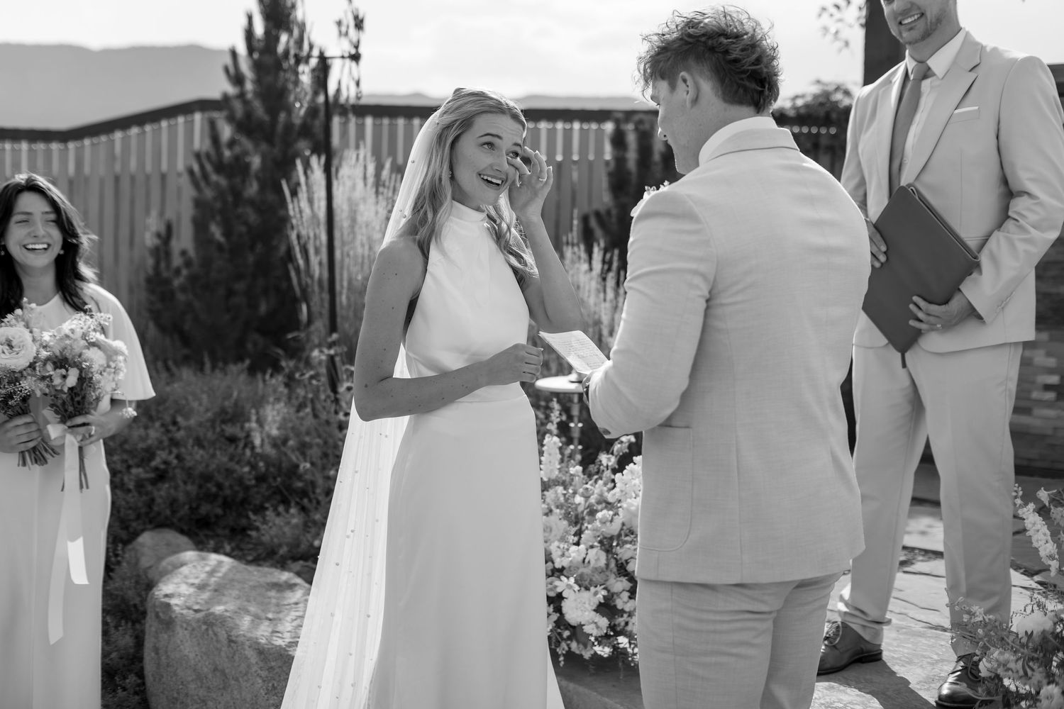 Black and white wedding ceremony photo of smiling bride and groom exchanging vows outdoors with officiant.