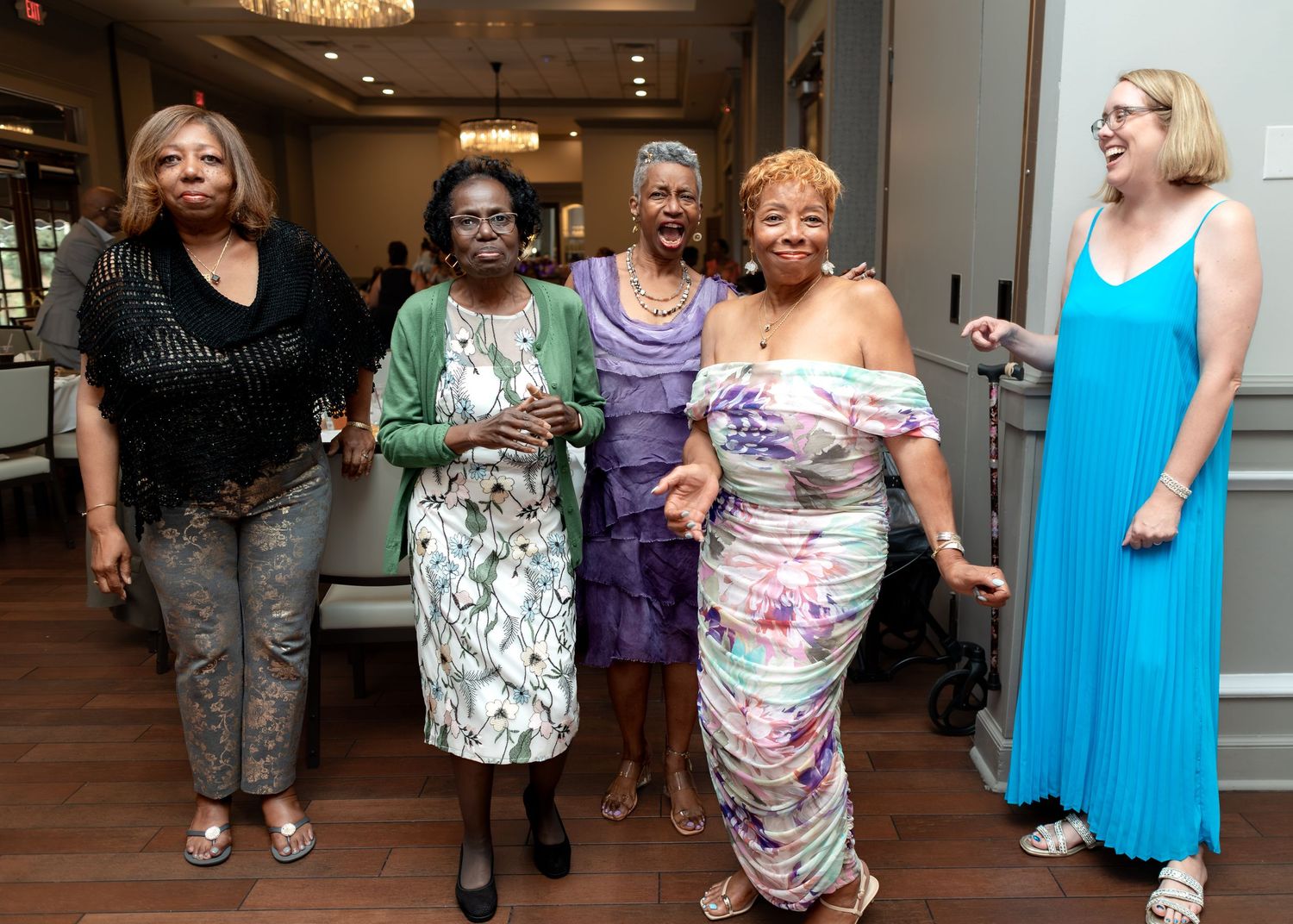 Five elegantly dressed women smile and pose together at a formal indoor celebration or party event.
