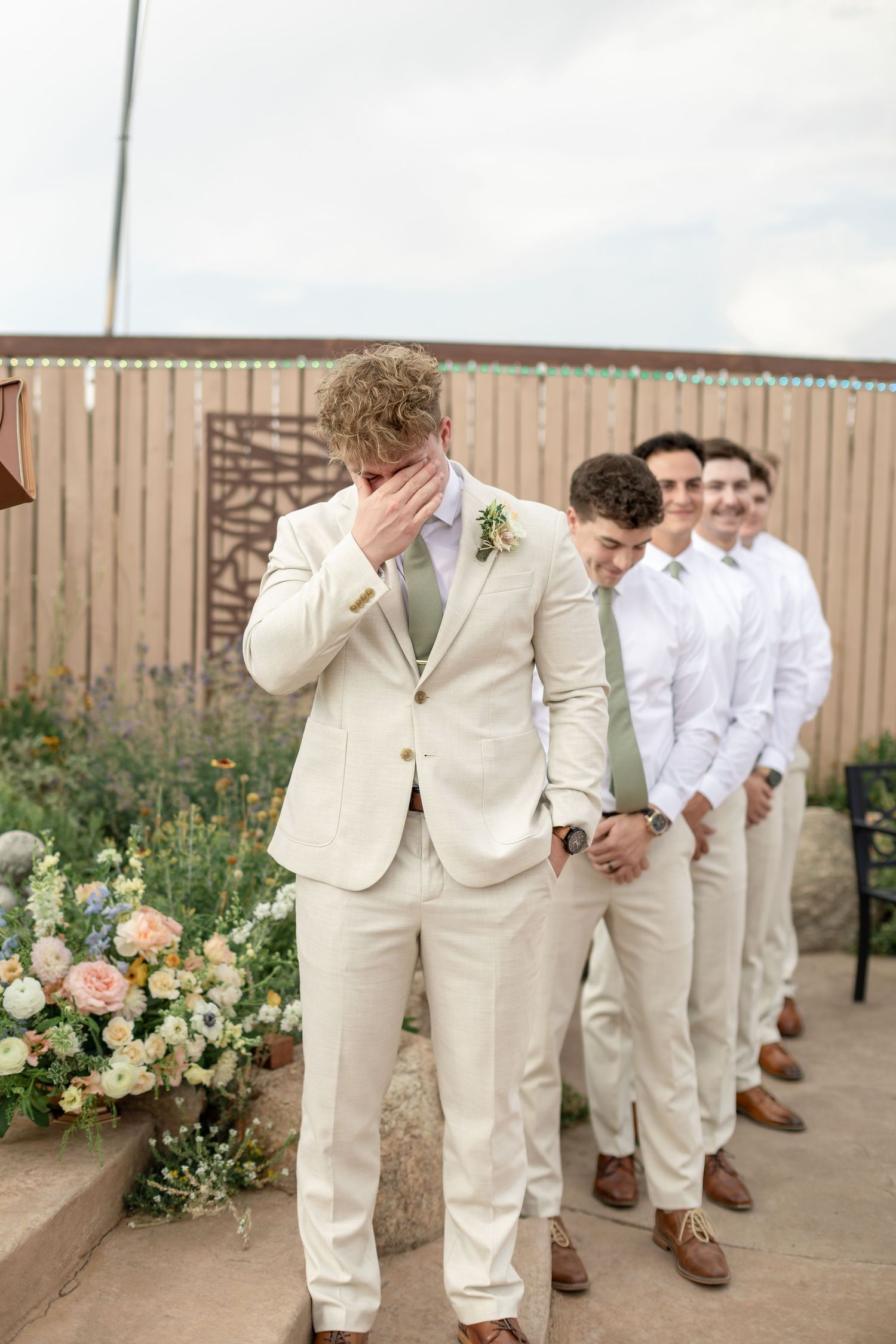 Emotional groom in cream suit covering his face as groomsmen line up behind him at outdoor wedding ceremony.