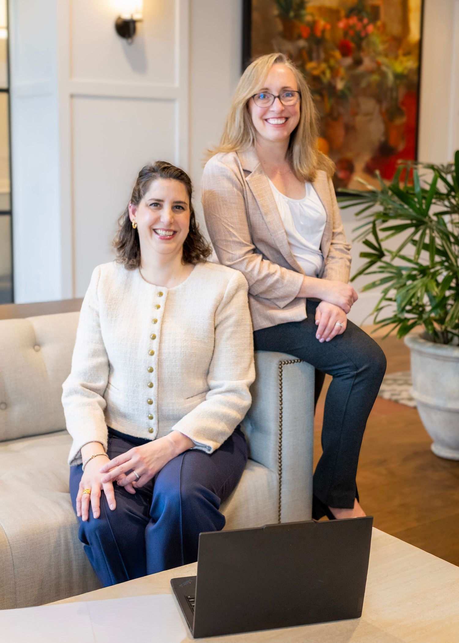 Professional seated in a gray office chair wearing a cream blazer and navy pants in a modern workspace setting.