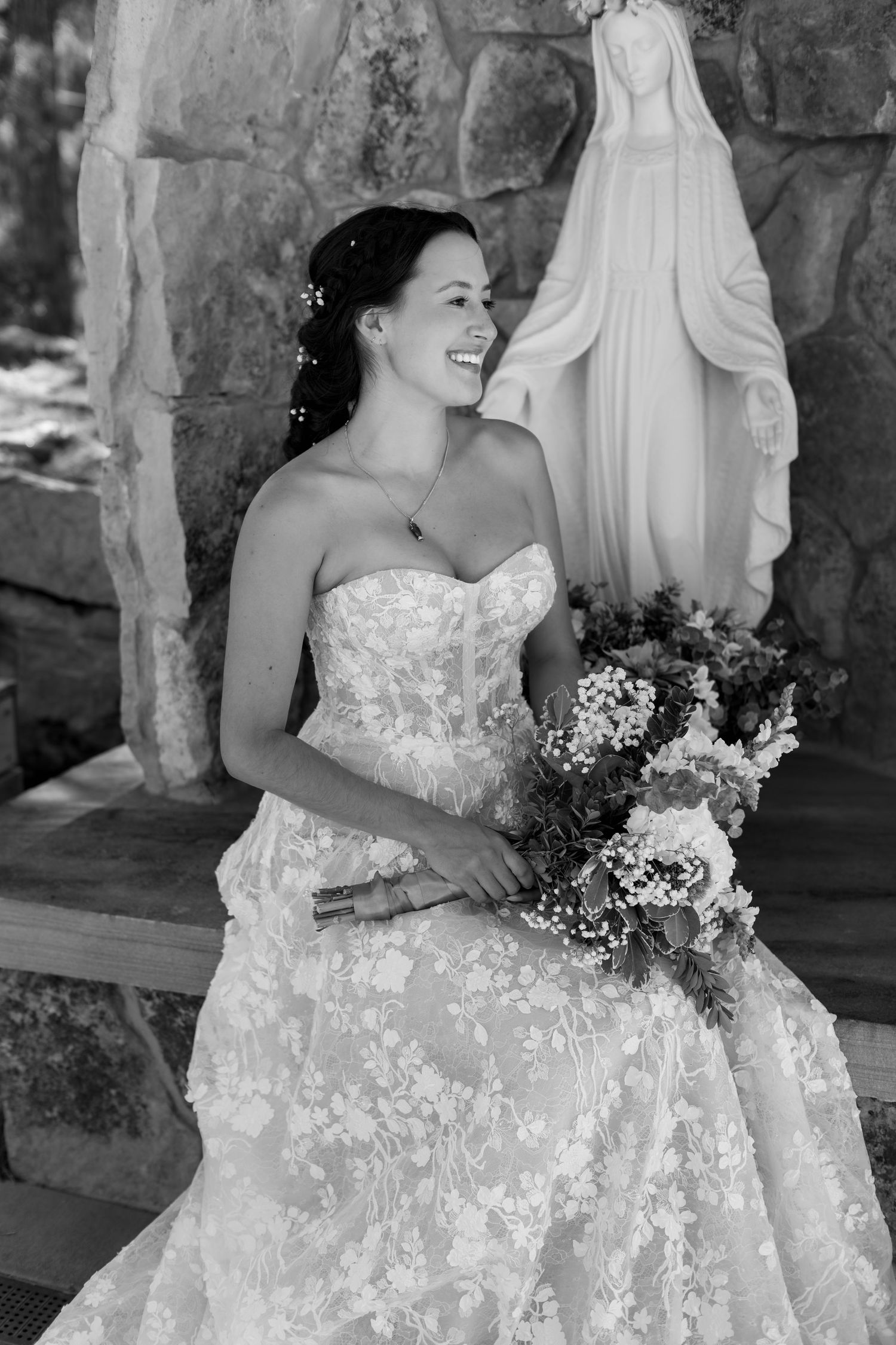Black and white photo of a smiling bride in lace gown holding bouquet beside a Virgin Mary statue.