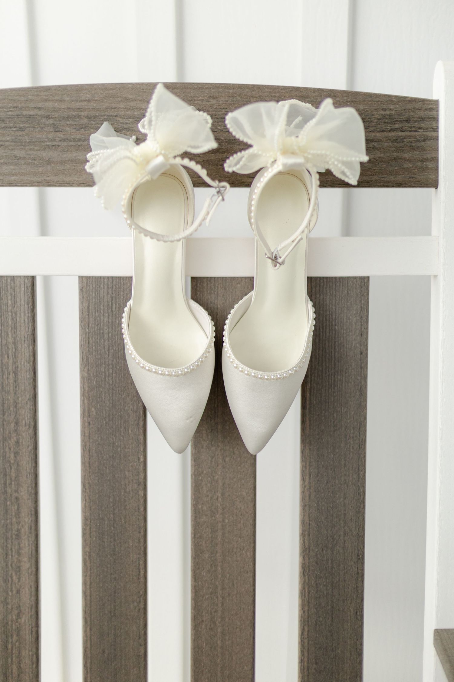 White bridal heels with pearl trim and organza bow ankle straps on wooden chair.