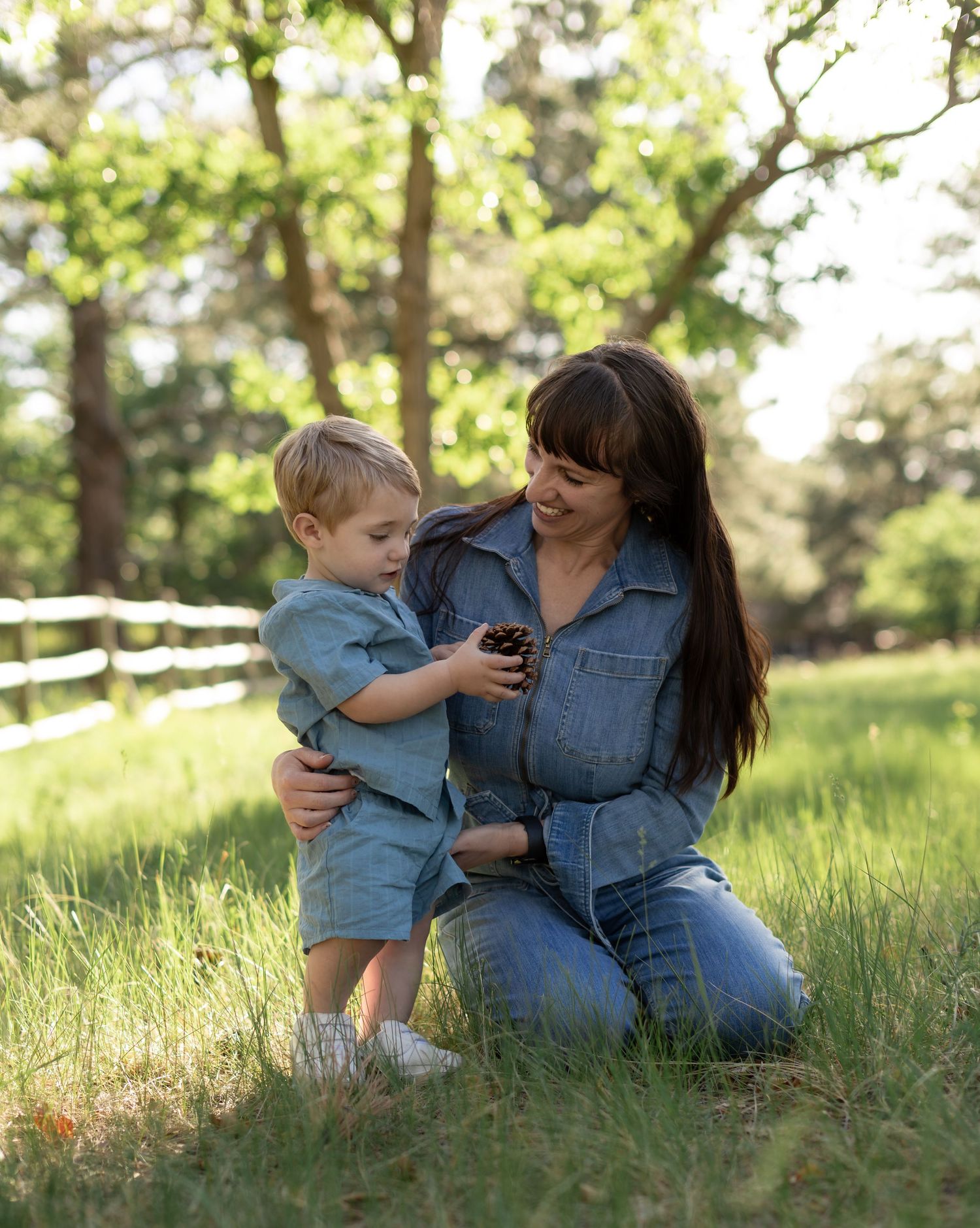 A woman in denim kneels on grass with a toddler boy examining a pinecone outdoors near a white fence.