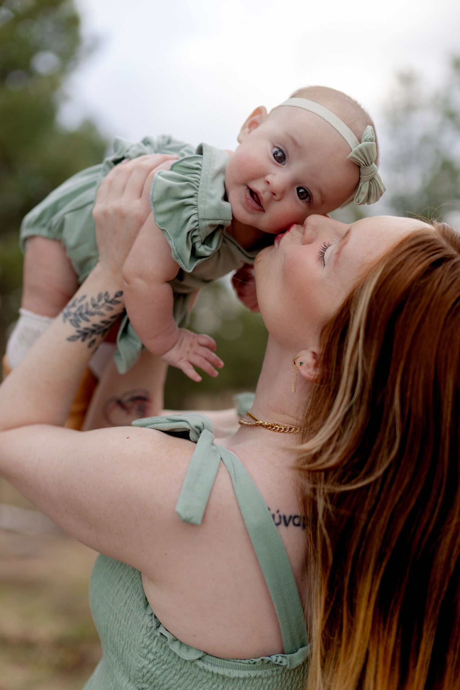 A redheaded mother lifts her smiling baby girl in a matching green outfit outdoors.