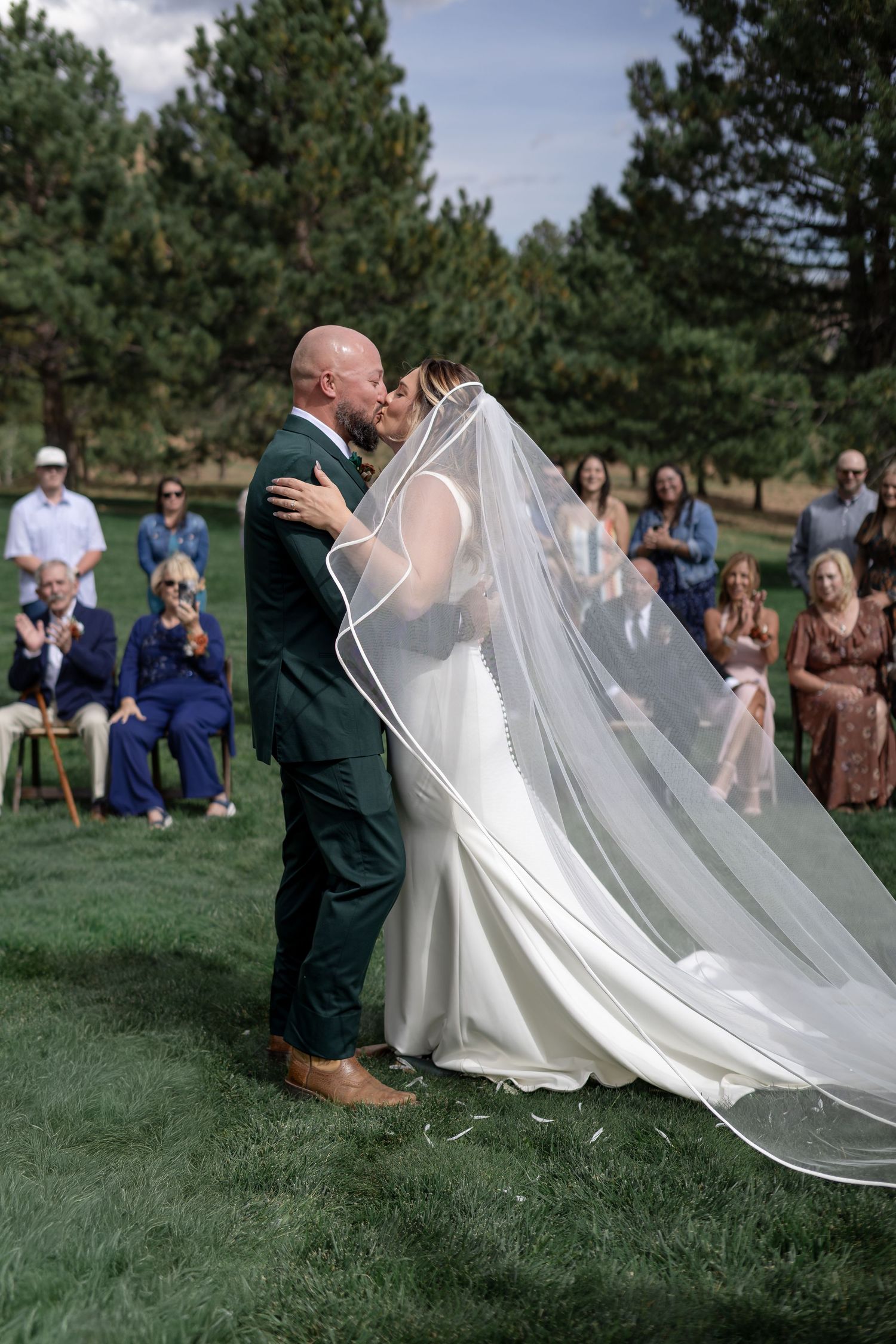 Bride and groom share first kiss at outdoor wedding ceremony with guests seated on grass surrounded by pine trees.