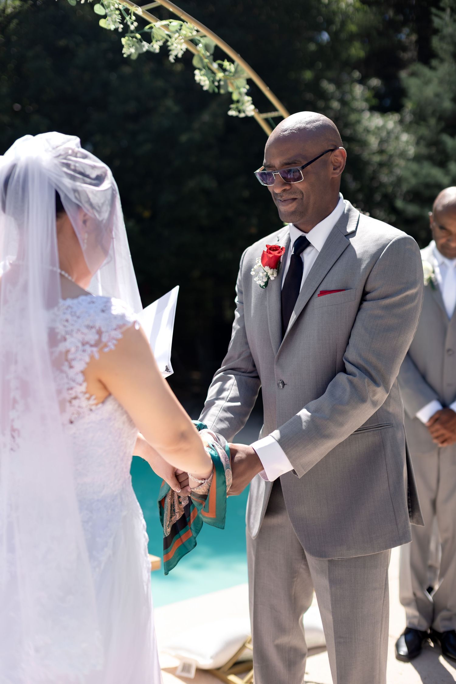 Groom in gray suit holds brides hands at outdoor wedding ceremony with floral arch backdrop.